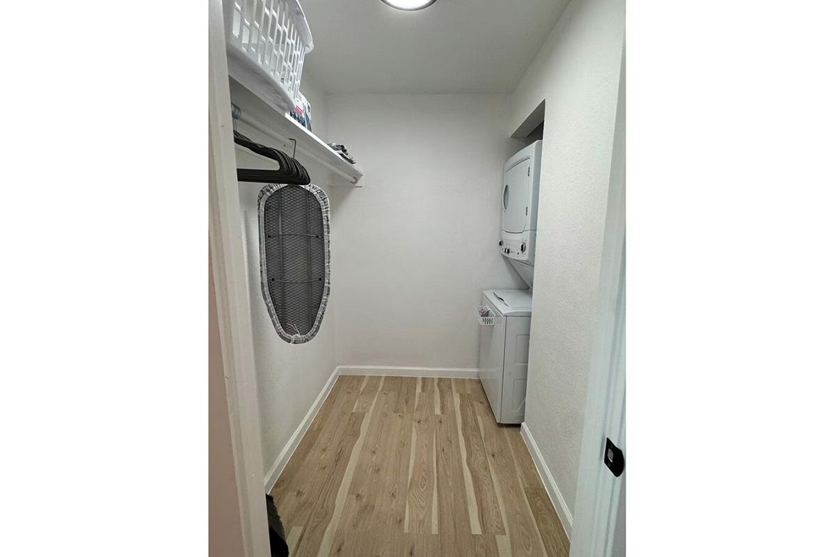 A small laundry room featuring a stackable washing machine and dryer, a white laundry basket on a shelf, and a hanging rod for clothes. The floor is made of light wood, and the walls are painted white, giving the space a clean and organized appearance.