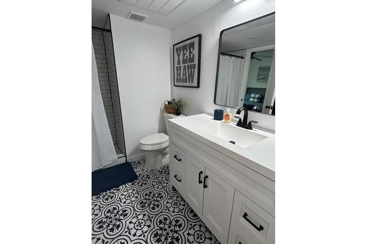 A clean, modern bathroom featuring a white vanity with a sink, a large mirror, and a stylish black and white patterned floor. There's a shower with a curtain, a small plant on the counter, and a decorative sign on the wall that reads "YEE HAW." A blue mat is placed in front of the toilet.