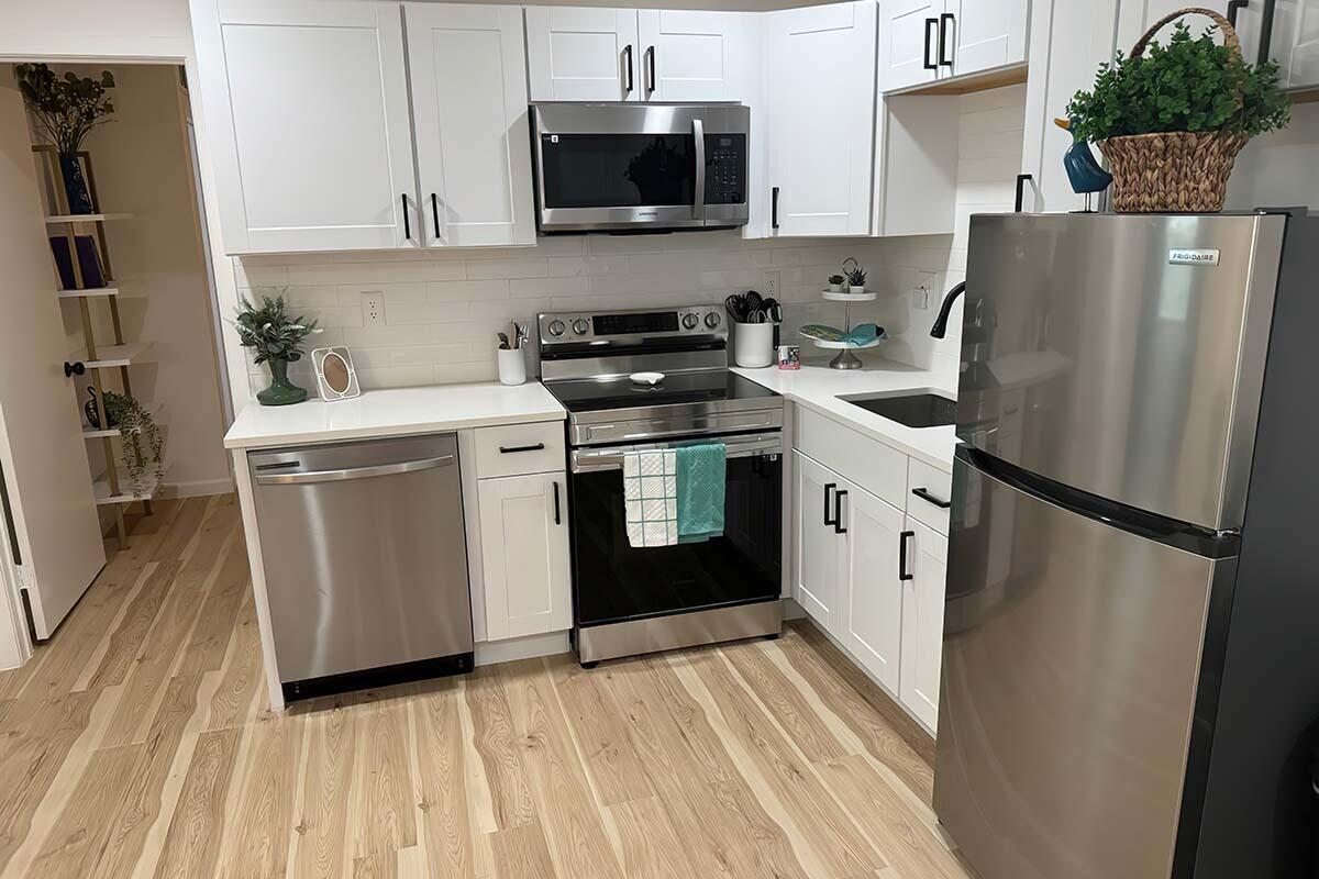 Modern kitchen featuring white cabinetry, stainless steel appliances including a refrigerator, microwave, dishwasher, and oven. A sink with a faucet is situated beside the stove. A decorative plant and dish towels add a touch of color. Light wood flooring enhances the airy, contemporary feel of the space.