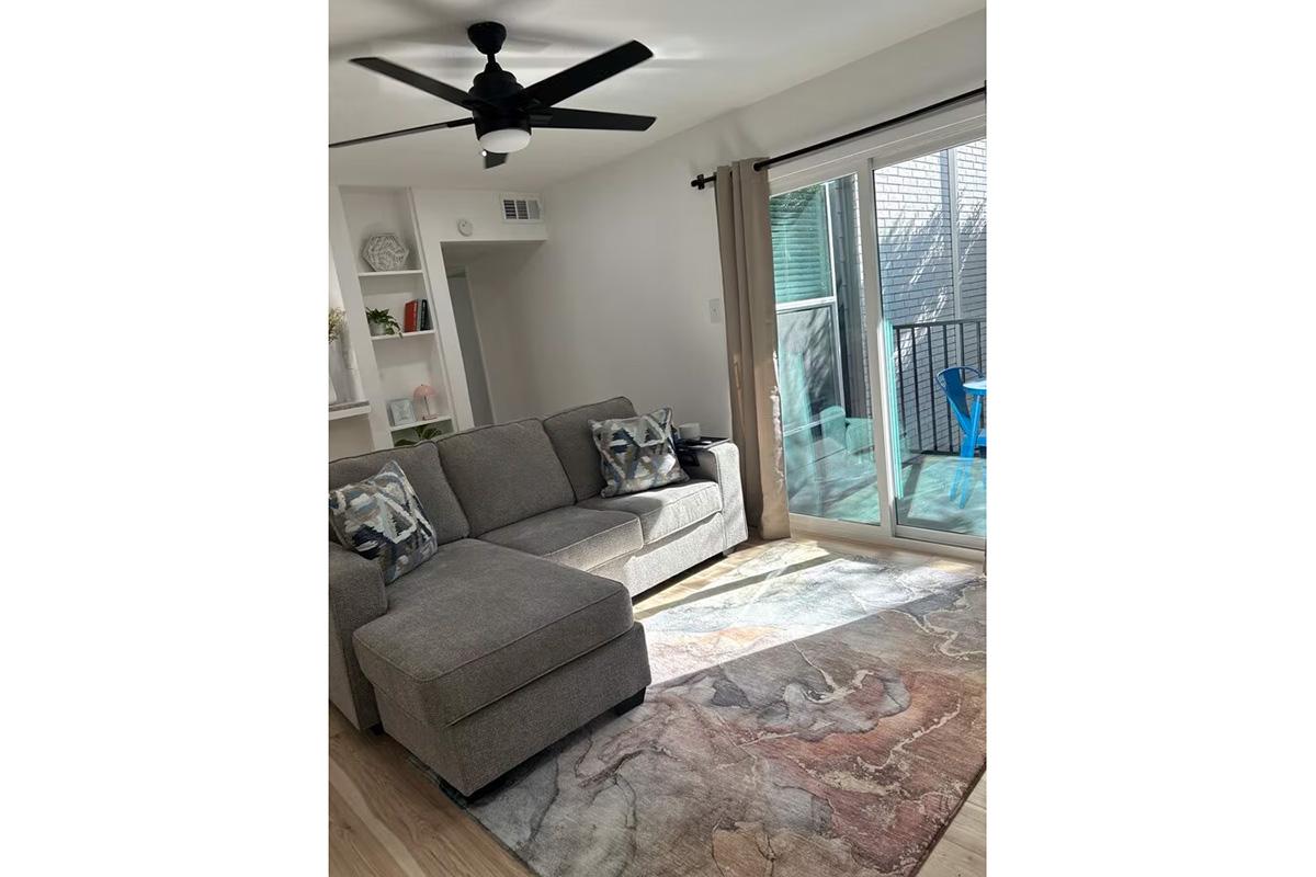 A cozy living room featuring a gray sectional sofa with decorative pillows, an area rug with a colorful design, and a ceiling fan. Large sliding glass doors open to a balcony with a blue chair visible outside. The space is well-lit and has a clean, modern aesthetic.