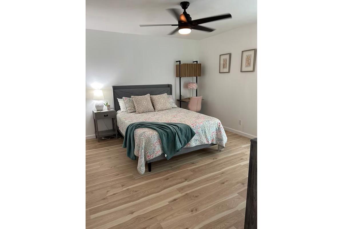 A cozy bedroom featuring a double bed with floral bedding, a nightstand with a lamp, and a modern ceiling fan. There are two framed artworks on the wall and a pink chair next to a small shelf, set against light wood flooring, creating a warm and inviting atmosphere.