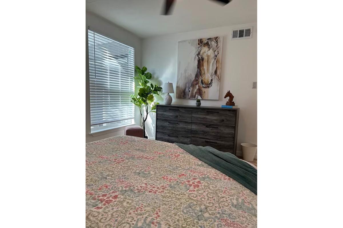 A cozy bedroom featuring a bed with a patterned quilt, a wooden dresser with a horse sculpture on top, and a large artwork of a horse on the wall. Natural light filters in through a window with blinds, complemented by a potted plant and a lamp to create a warm, inviting atmosphere.