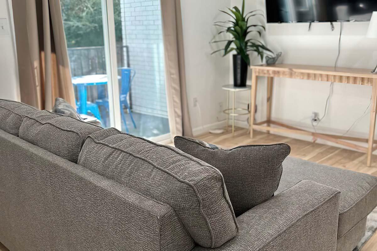 Cozy living room featuring a gray sofa with decorative pillows, a modern wooden console table, and a flat-screen TV mounted on the wall. A potted plant adds greenery, and a sliding glass door reveals an outdoor space with blue chairs. Natural light streams in, enhancing the inviting atmosphere.