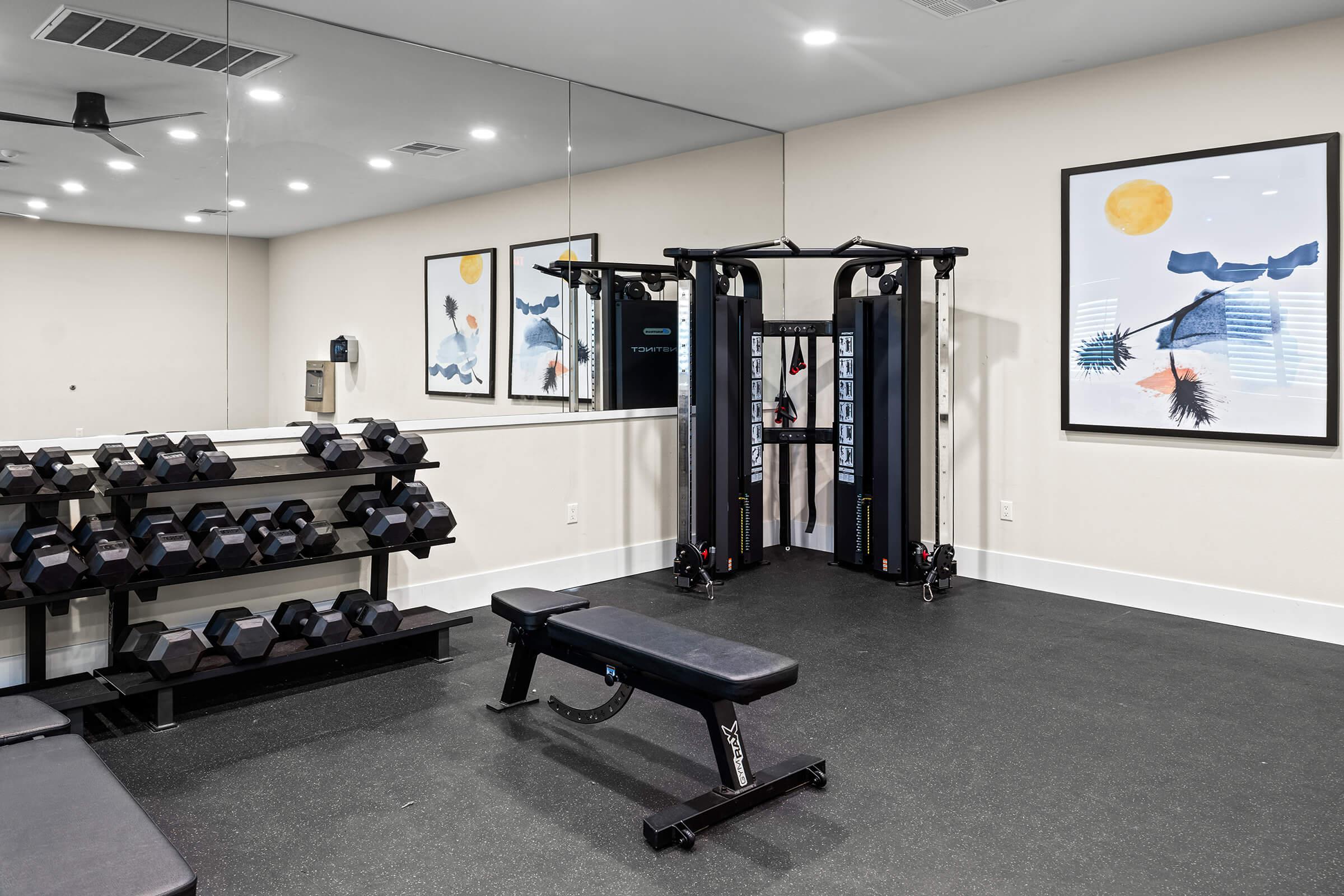 A gym featuring a workout area with a bench, a rack of dumbbells, and an exercise machine. There is a large mirror. 