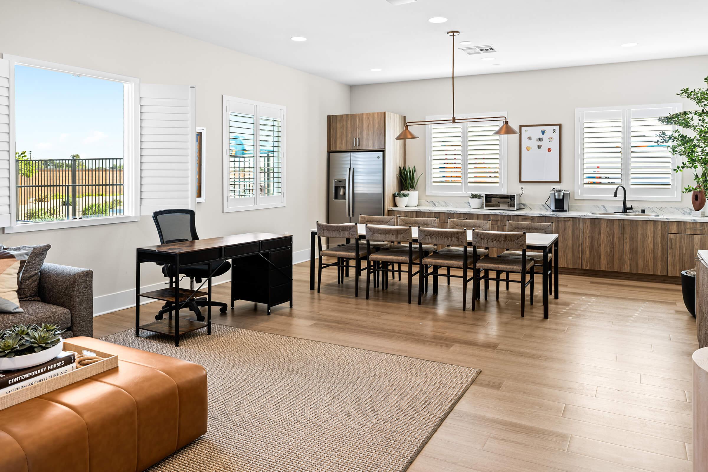 An open-concept living space with a seating area, a desk with a chair, and a dining table with chairs. The kitchen is stylish, and large windows provide natural light. 