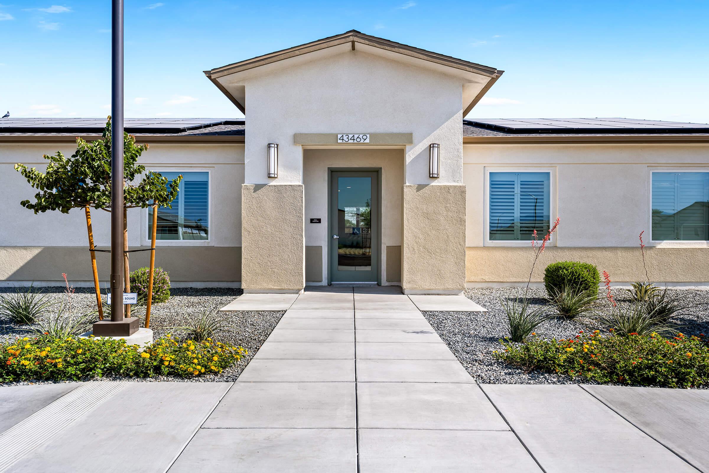 A single-story building with a light-colored facade. The entrance has a glass door framed by a small porch, with a landscaped area with shrubs, flowers, and decorative stones. 