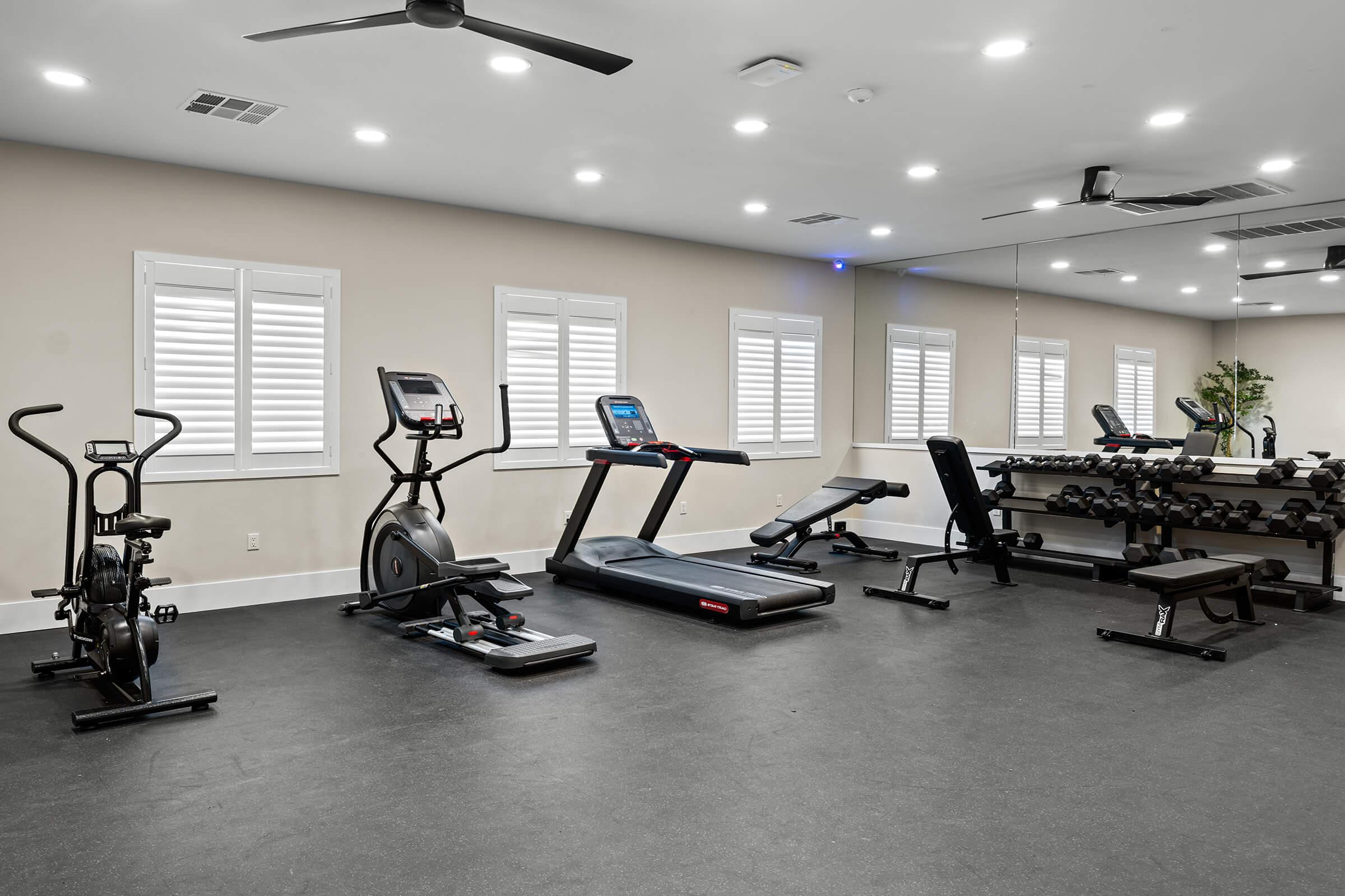 A gym with various fitness equipment. There is an elliptical machine, a treadmill, a weight bench, and a set of dumbbells. 