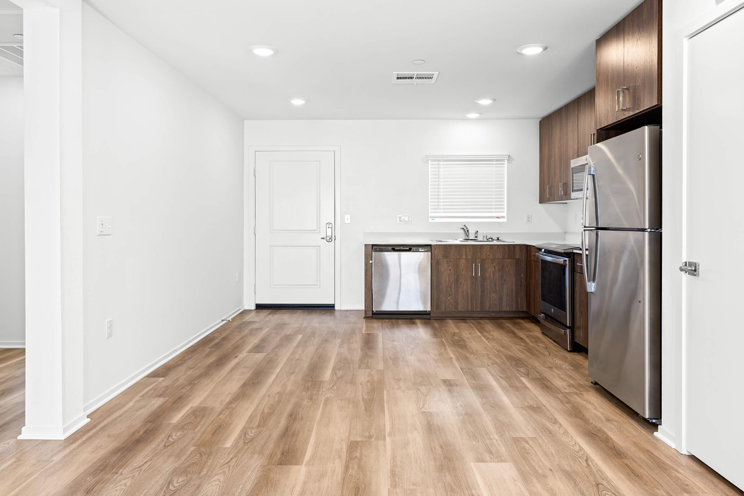 A kitchen with stainless steel appliances, including a refrigerator and dishwasher. There are wooden cabinets. Light-colored walls and wooden flooring. 