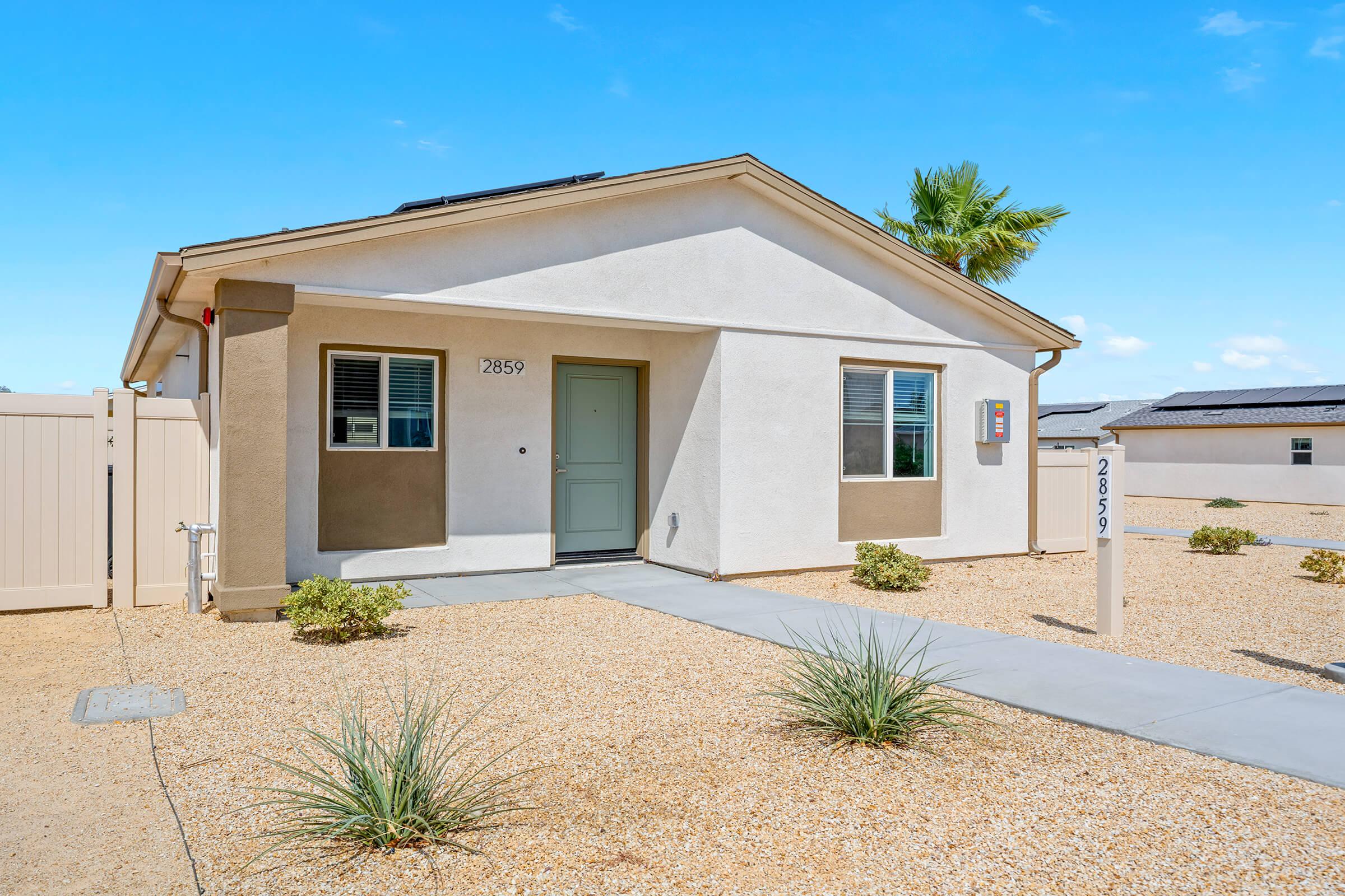 A single-story house with a light-colored exterior and a front entryway, surrounded by gravel landscaping and small plants. 