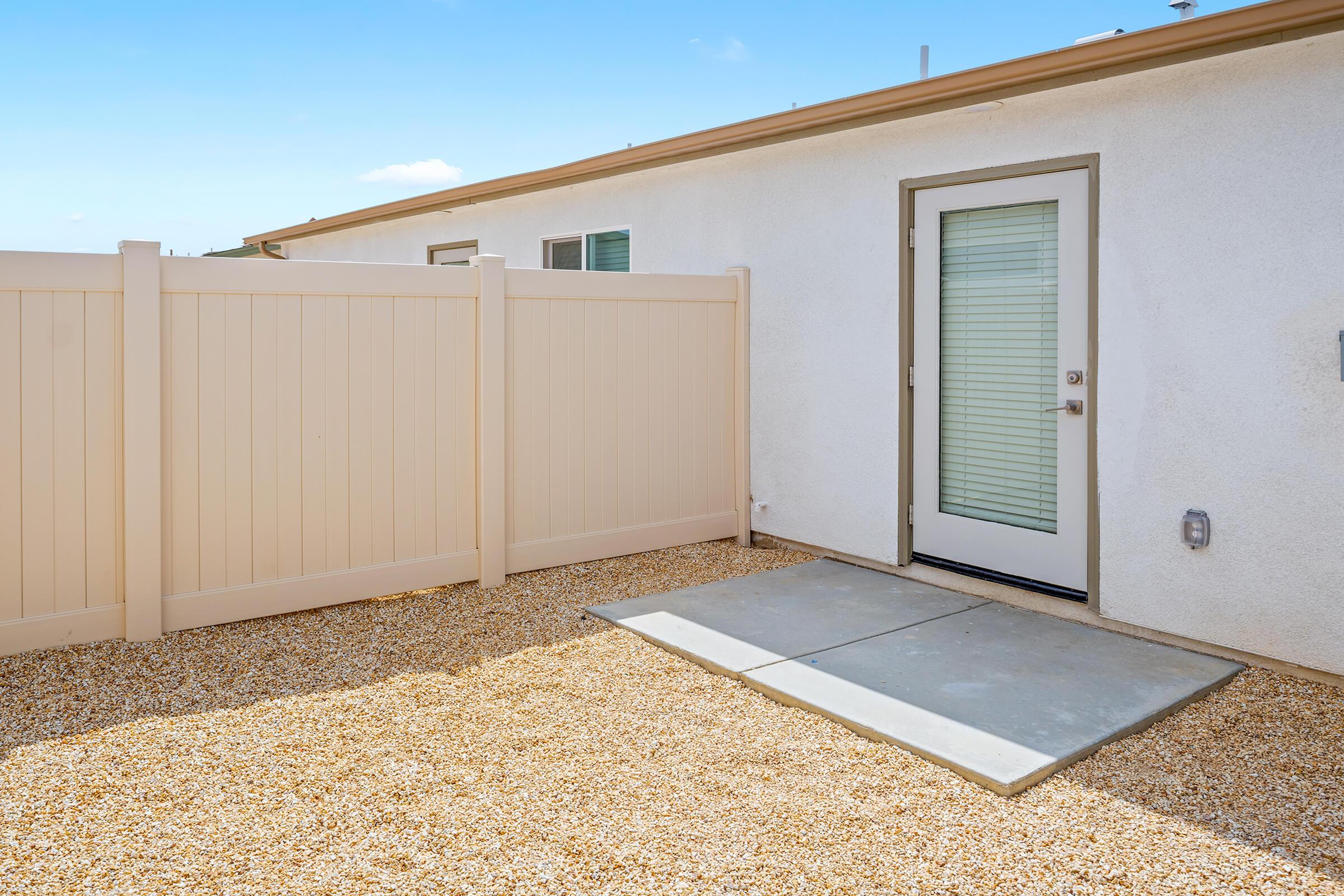 A small outdoor area with light-colored gravel, enclosed by a beige privacy fence under a clear blue sky.