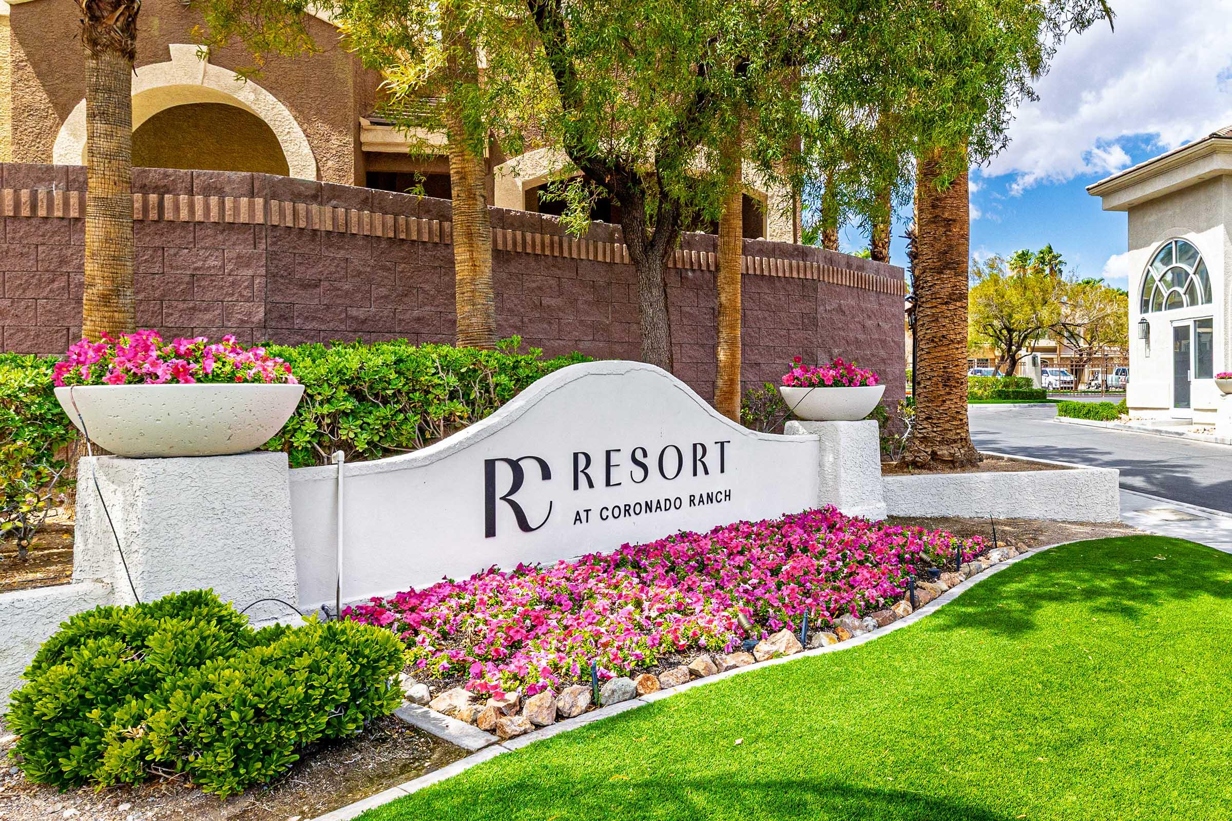 Sign for "R Resort at Coronado Ranch" surrounded by colorful flowers and well-maintained landscaping. The building is partially visible in the background, with palm trees adding to the tropical aesthetic. The scene is bright and welcoming, suggesting a pleasant resort environment.