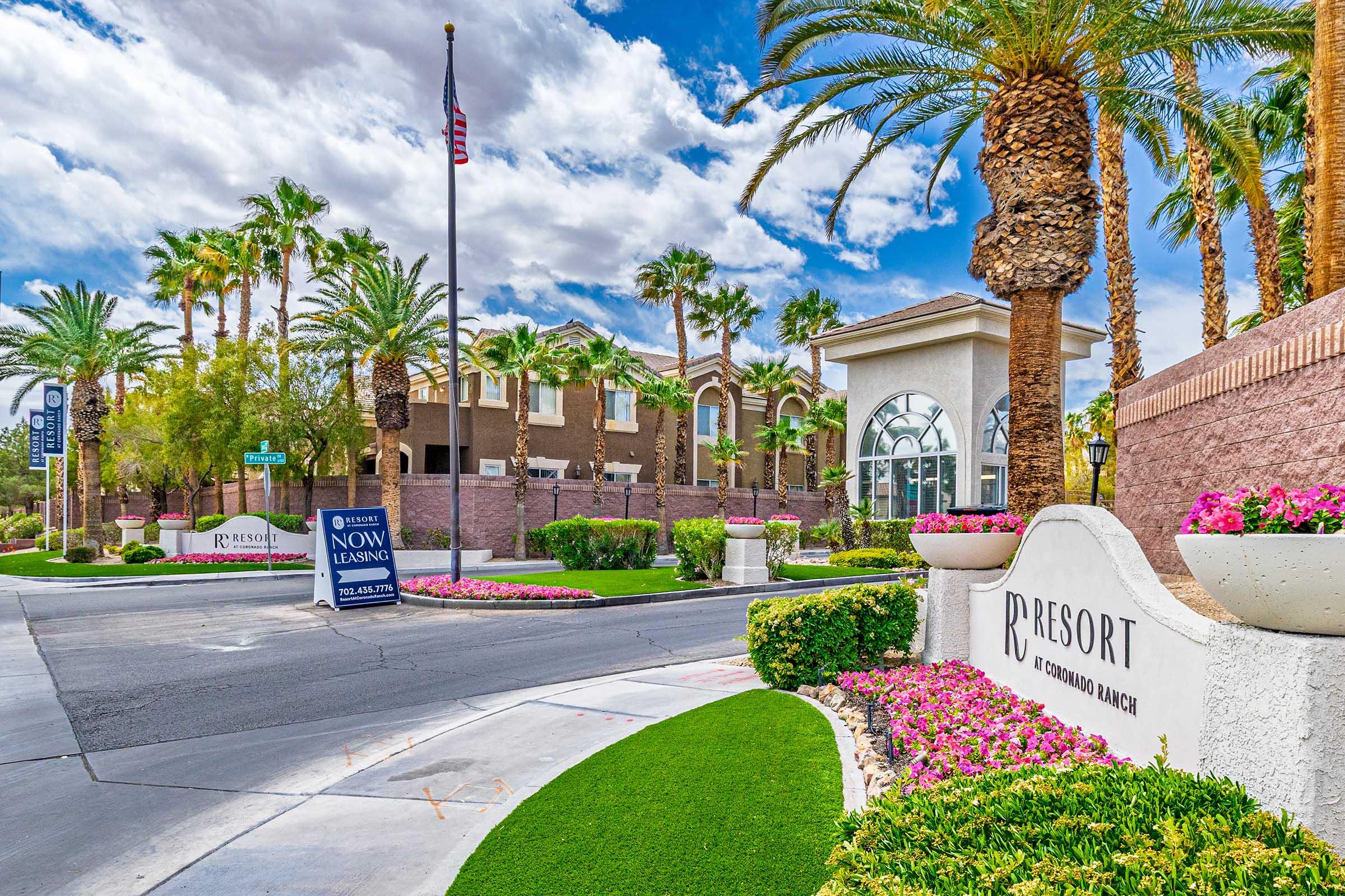 Entrance to a resort with a sign that reads "R Resort." Palm trees line the driveway, and colorful flowers are planted along the path. The sky is partly cloudy, and a flag stands prominently in the background. Signs indicate leasing and real estate opportunities nearby.