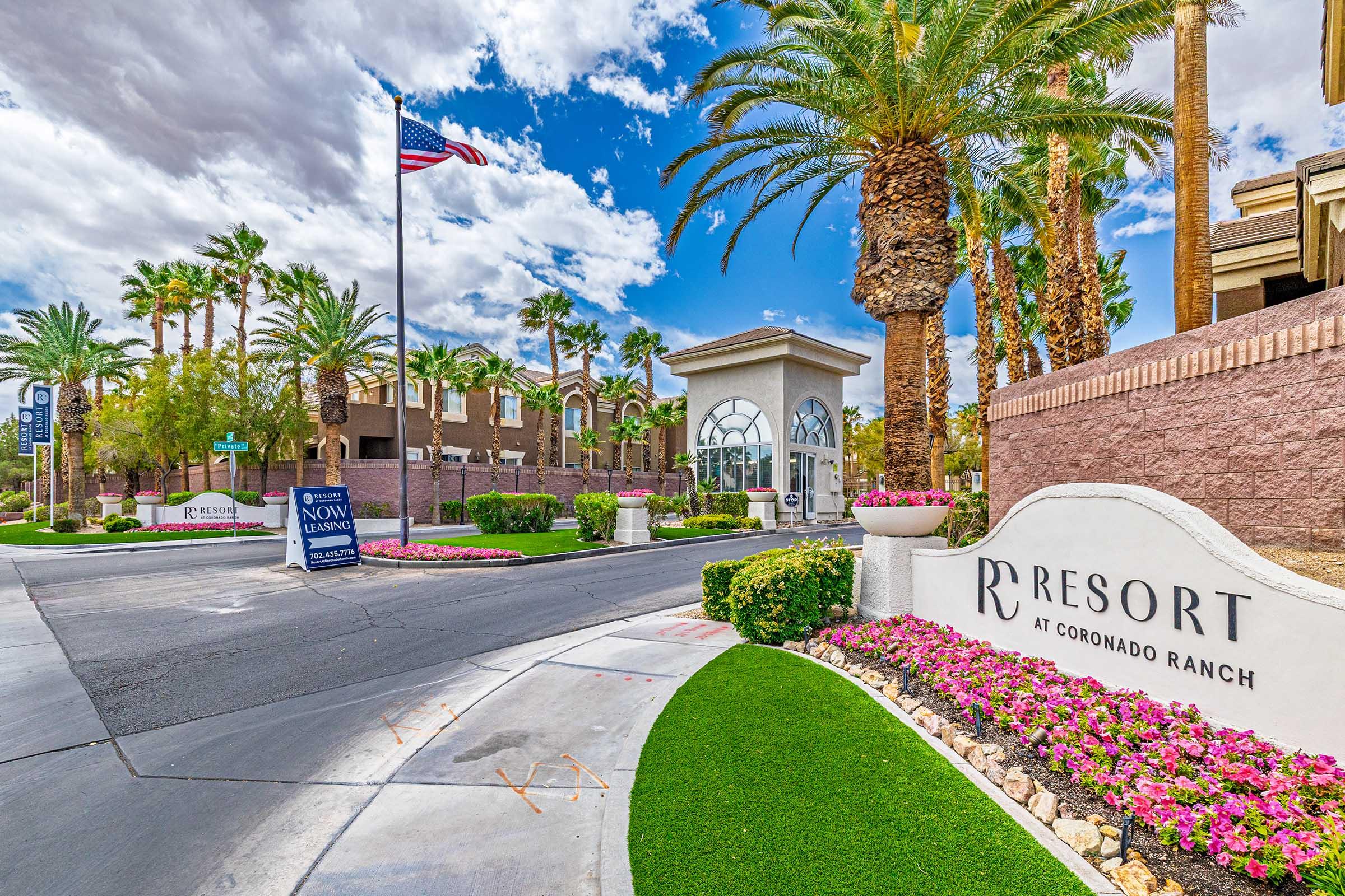 Entrance to the Resort at Coronado Ranch featuring palm trees, colorful flower beds, and a sign. An American flag is visible in the background under a partly cloudy sky. The pathway is lined with greenery, creating a welcoming atmosphere.