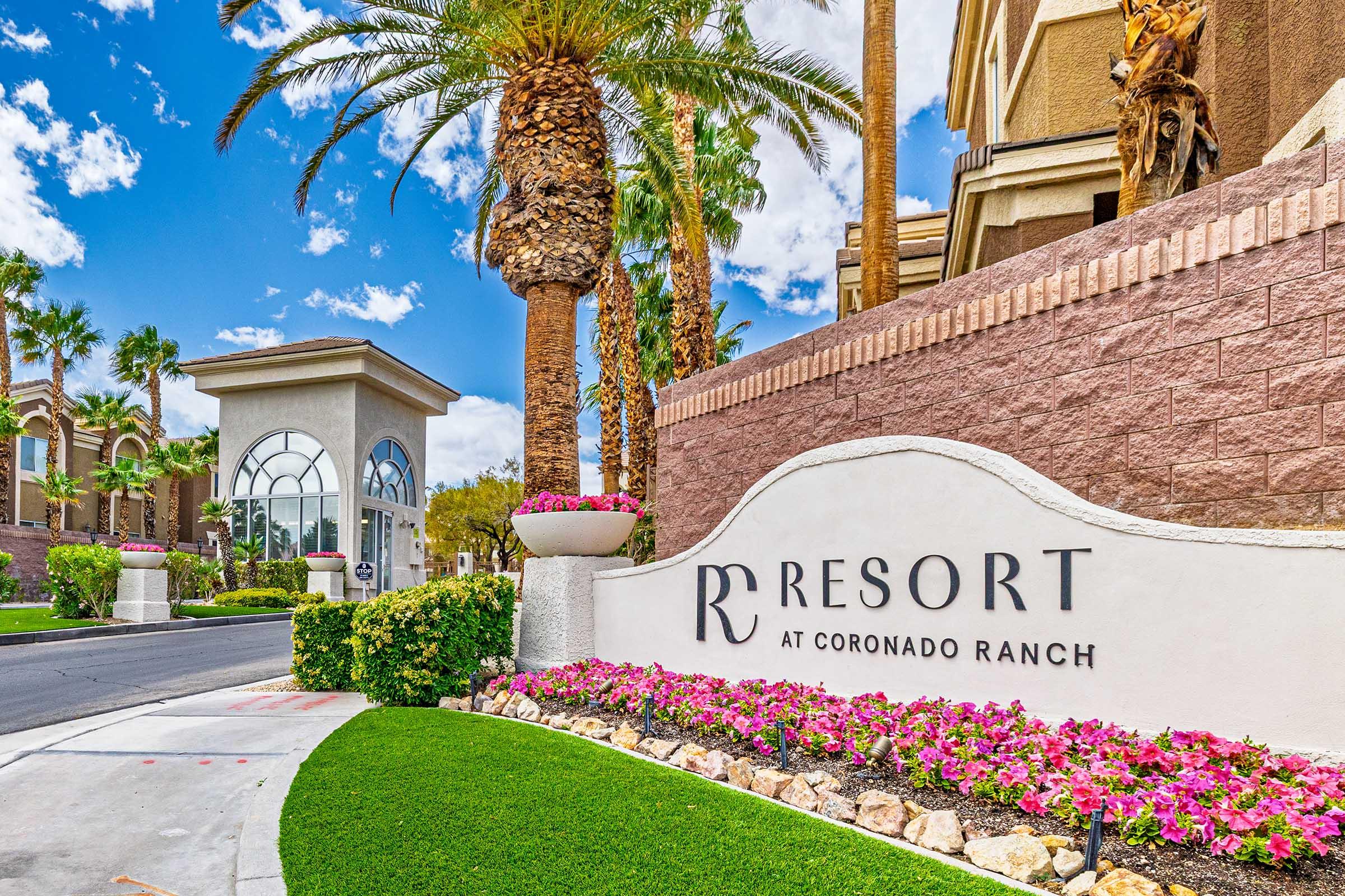 Sign for the Resort at Coronado Ranch, featuring elegant typography. The setting includes manicured greenery, colorful flower beds, and tall palm trees under a bright blue sky with fluffy clouds. A gateway structure is visible in the background, adding to the inviting atmosphere of the resort entrance.