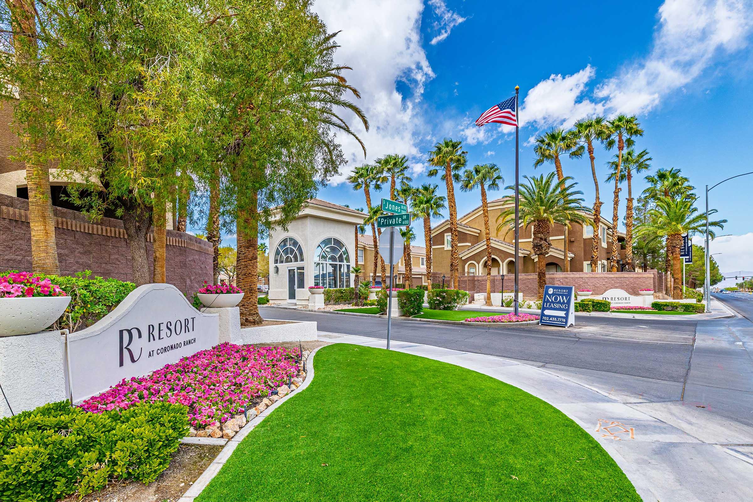 A well-maintained entrance to a resort, featuring a sign that reads "RV Resort," surrounded by colorful flower beds and palm trees. An American flag is flying in the background, and there are directional signs indicating various attractions nearby. The sky is bright blue with a few clouds.
