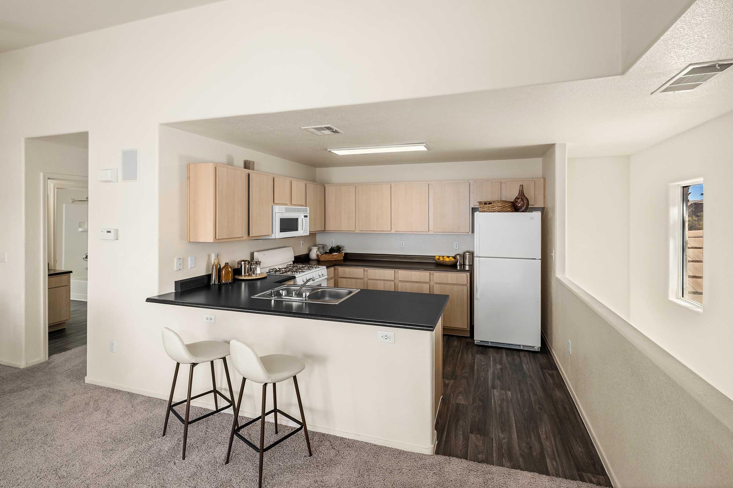 Modern kitchen with light wood cabinetry, a black countertop, and stainless steel appliances. Features include a microwave, white refrigerator, and a small breakfast bar with two white stools. The space is well-lit and has neutral-colored walls and carpeting.