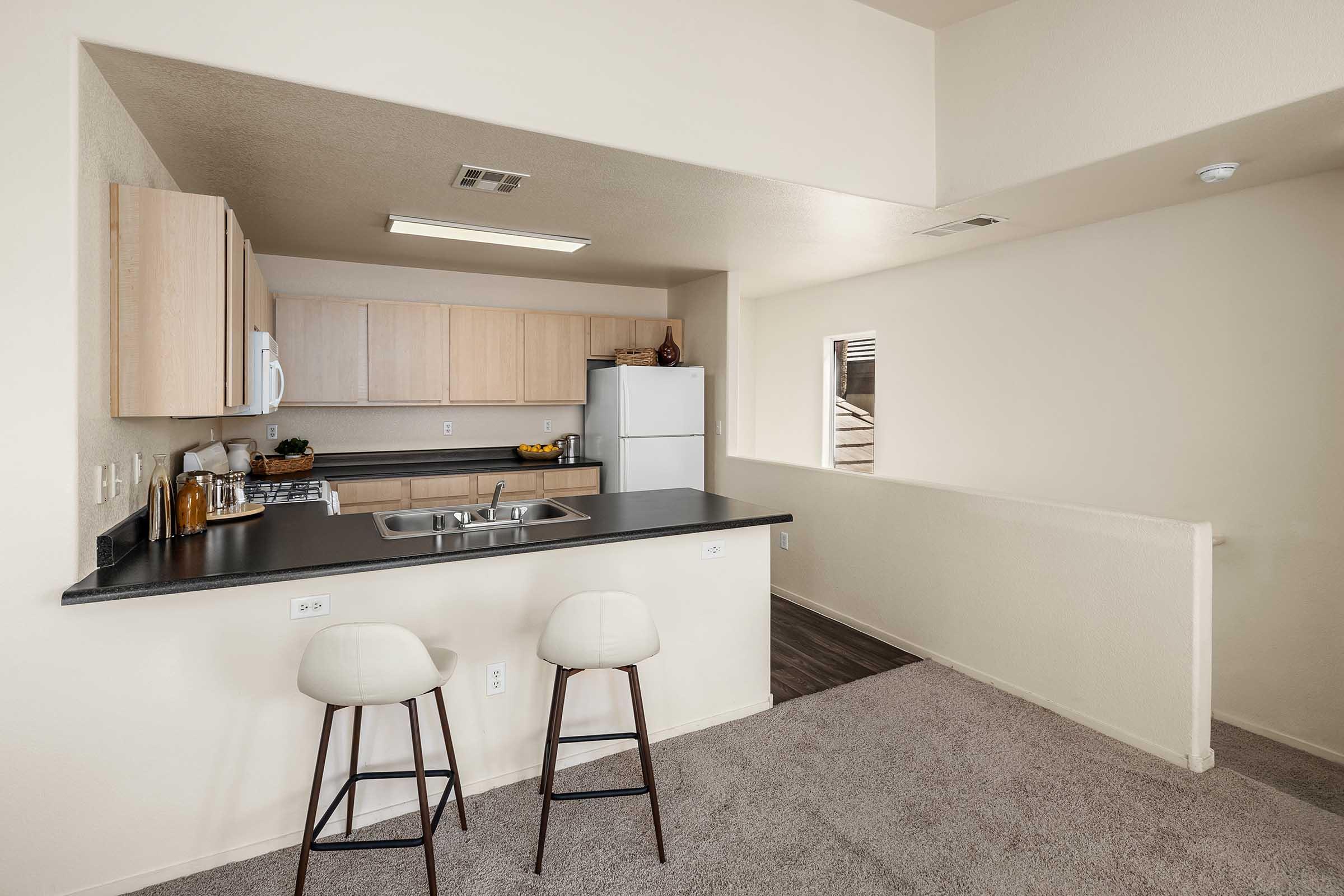 A modern kitchen space featuring light wood cabinetry, a white refrigerator, and a dark countertop. Two beige bar stools are positioned at the kitchen island. The walls are painted in neutral tones, and there are minimal decorative items. A window provides natural light to the area.