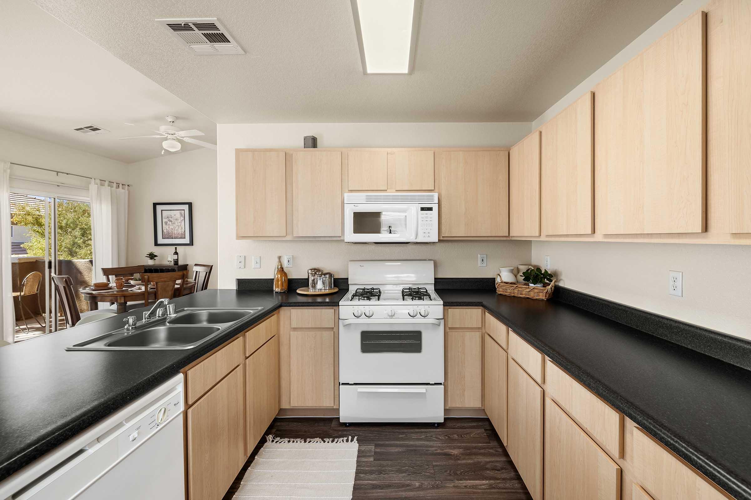 A modern kitchen featuring light wood cabinets, a black countertop, a white stove, and a microwave. There is a sink with a double basin, a dishwasher, and a small dining area visible in the background. Natural light enters through a window, enhancing the open and inviting atmosphere.