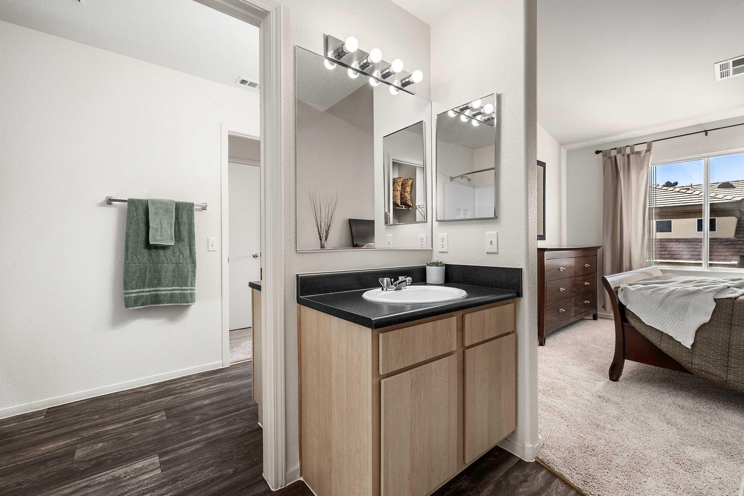 A modern bathroom featuring a double sink vanity with a dark countertop, two mirrors, and stylish lighting. There is a towel rack with a green towel, and a doorway leading to another room. In the background, a bedroom is visible with a bed and dresser, and large windows offering natural light.