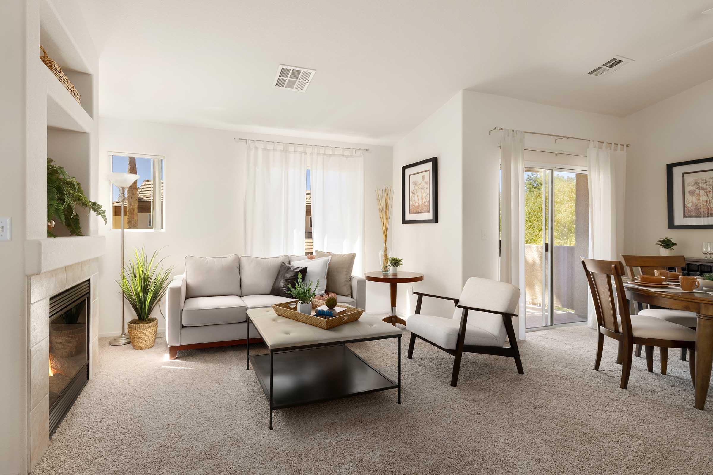 A stylish living room featuring a light gray sectional sofa, a coffee table, and a comfortable armchair. Natural light fills the space, highlighting a decorative plant near a stone fireplace. The area includes a circular dining table and a sliding glass door leading to an outdoor terrace.