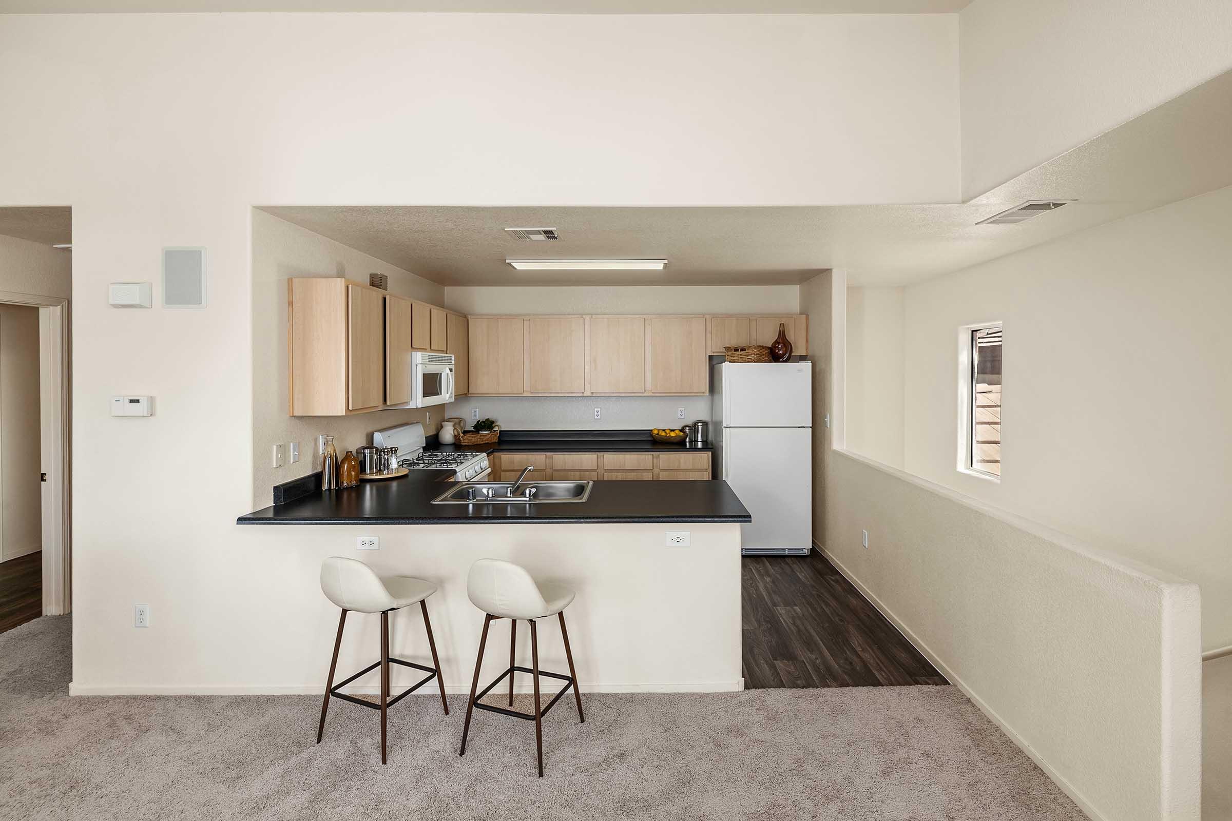 A modern kitchen with light wood cabinetry, a white refrigerator, and a white microwave. There is a black countertop with two bar stools, and the kitchen is well-lit with natural light coming from a nearby window. The flooring is a light-colored carpet, creating a cozy atmosphere.