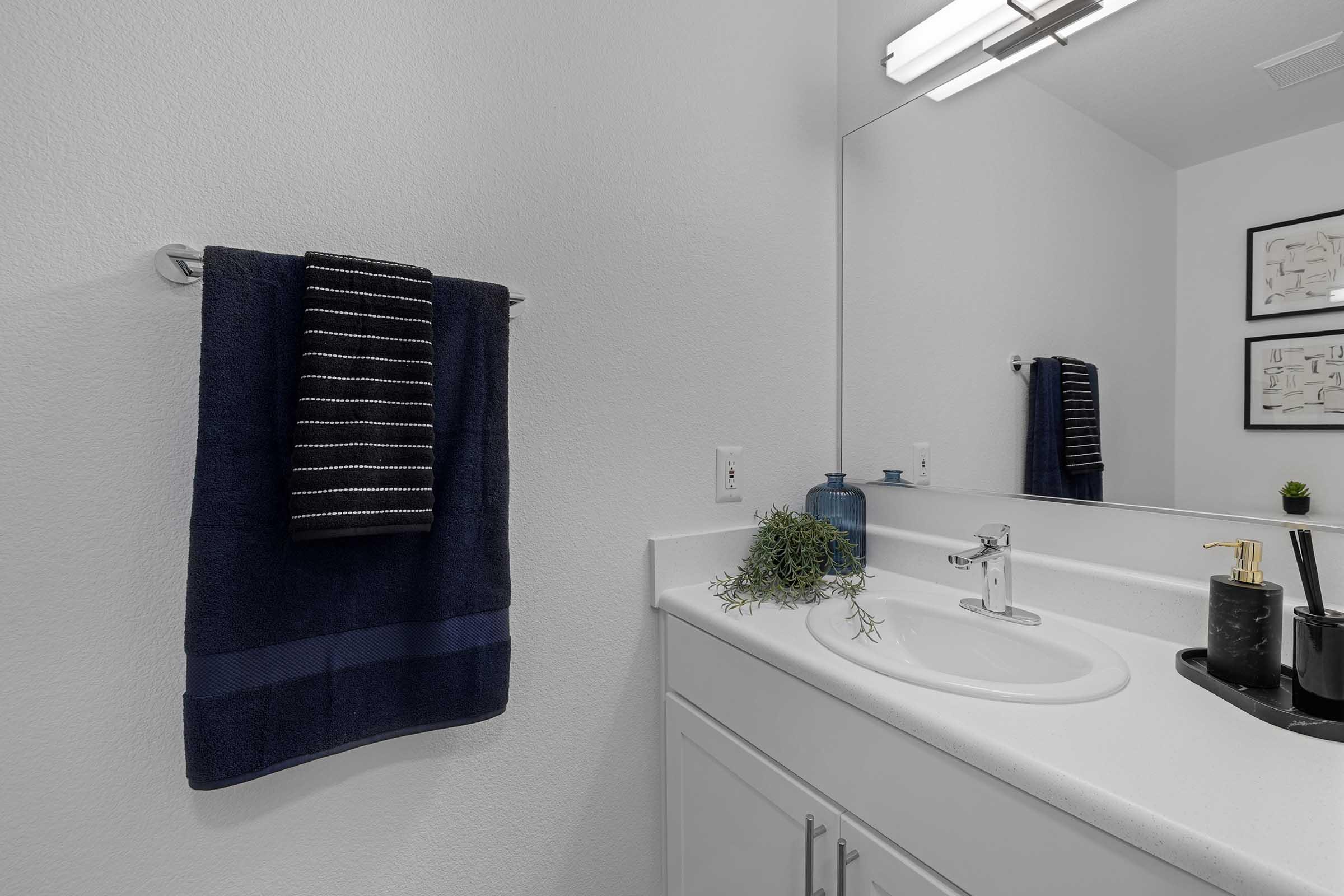 A modern bathroom featuring a white sink and countertop, a large mirror, and wall-mounted towel rack with a dark striped towel. Decorative elements include a small green plant and blue vase on the counter, with minimalistic framed art on the wall. The color palette is neutral with hints of dark blue.