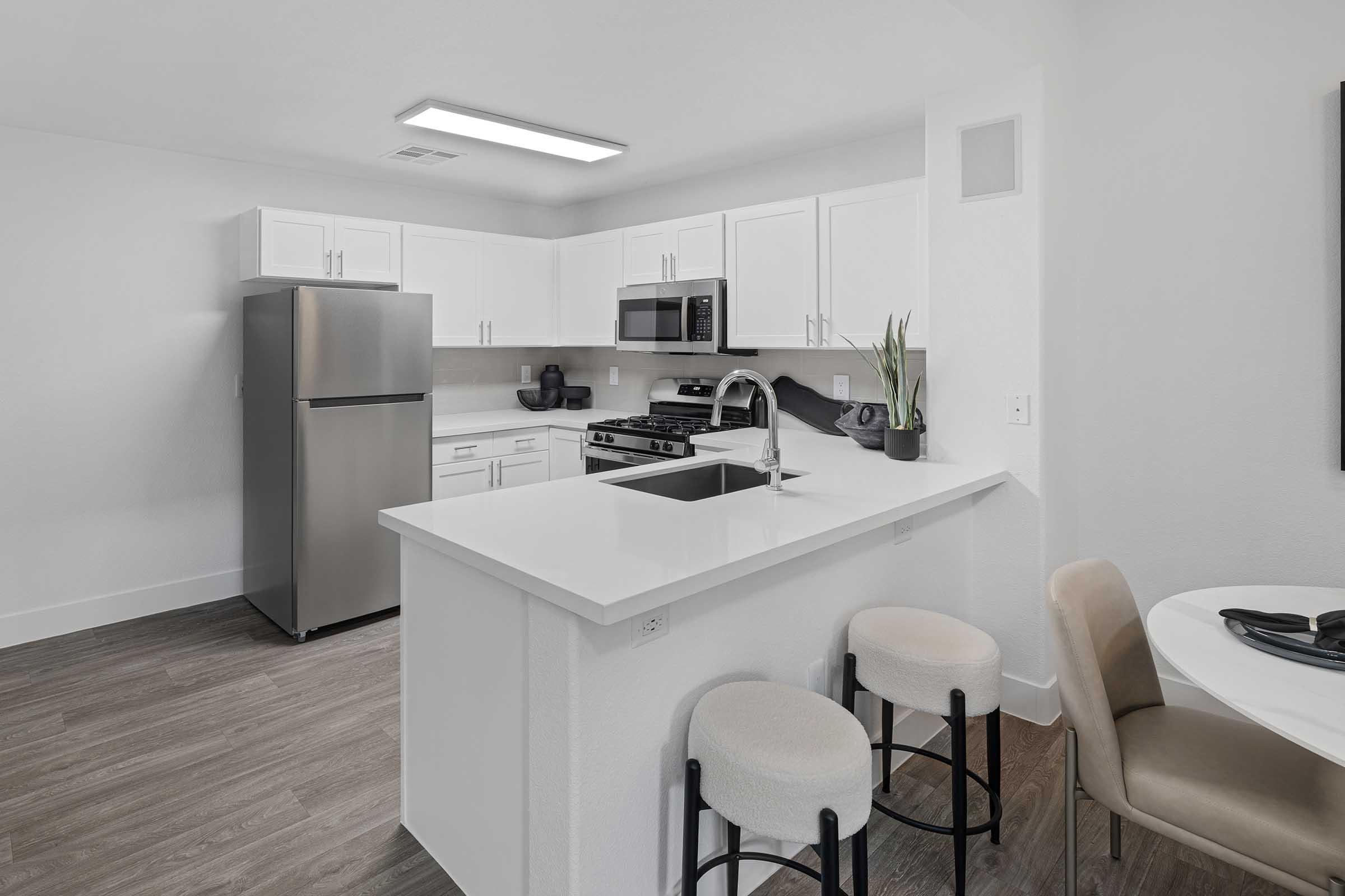 Modern kitchen featuring white cabinetry, stainless steel appliances including a fridge and microwave, a white island with a sink, and two beige bar stools. The space has a clean, minimalist design with light wood flooring and a small dining area visible in the background.