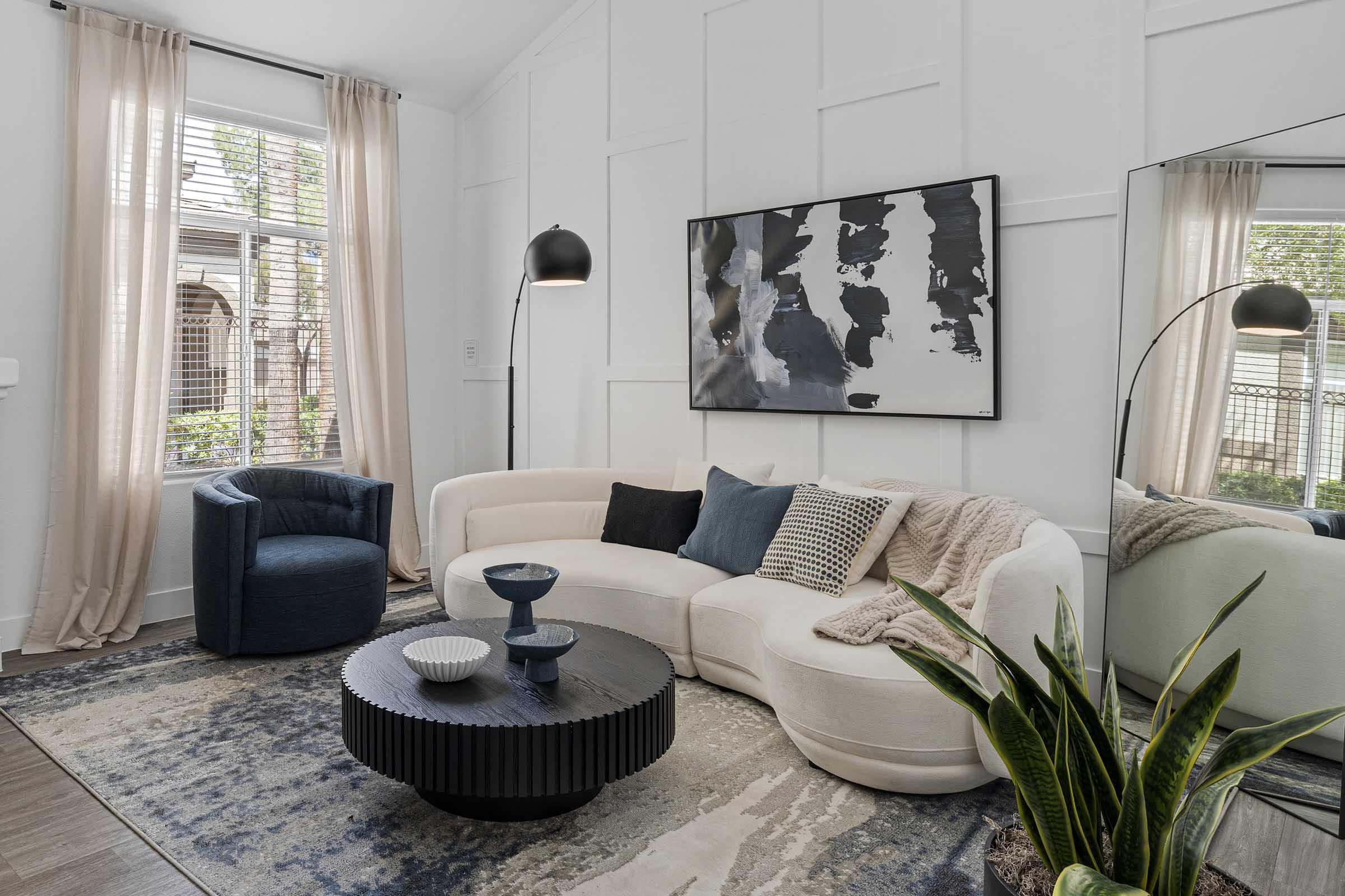 A modern living room featuring a sleek, white curved sofa with decorative pillows, a round black coffee table, and a textured area rug. A tall floor lamp provides soft lighting, while a chair in deep blue complements the decor. Plants and a large mirror add to the space's stylish ambiance. Natural light filters through sheer curtains.