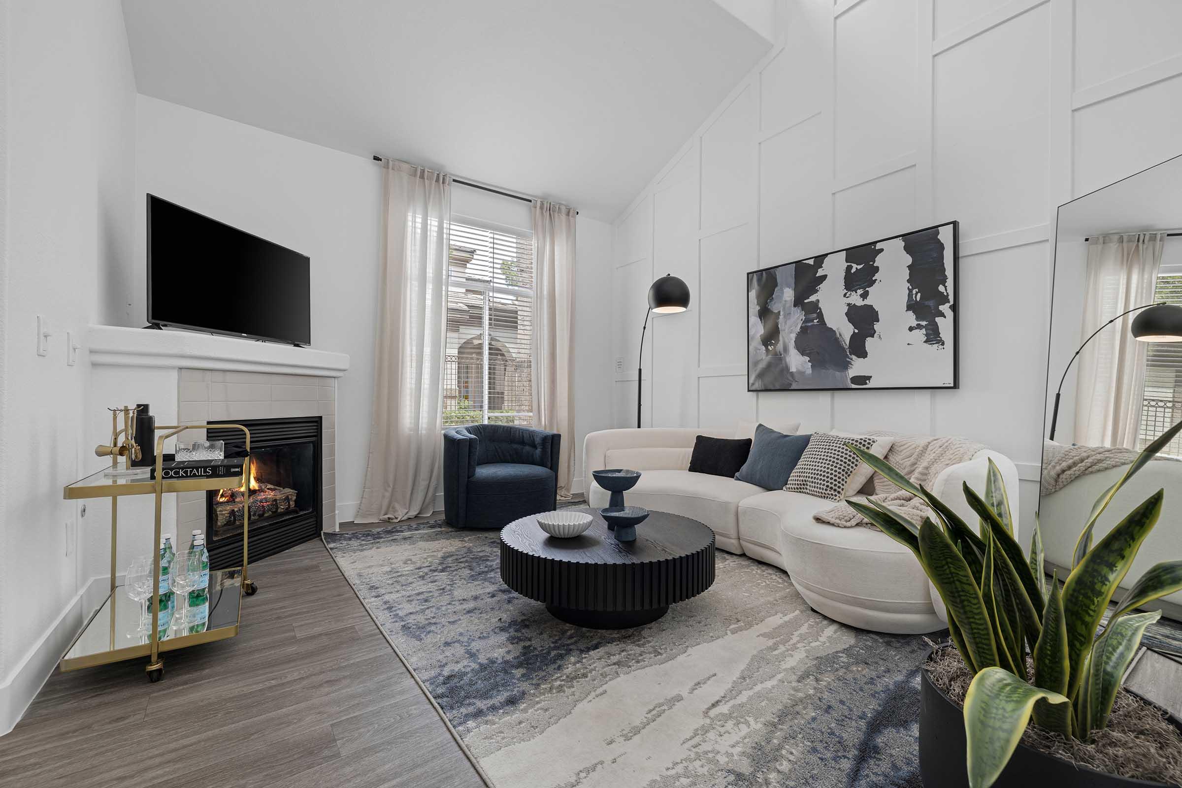 Modern living room featuring a white sectional sofa, a round black coffee table, and a stylish floor lamp. A flat-screen TV is mounted above a fireplace. Large windows with sheer curtains allow natural light, while a potted plant adds greenery. The decor includes abstract artwork and a bar cart with drinks.