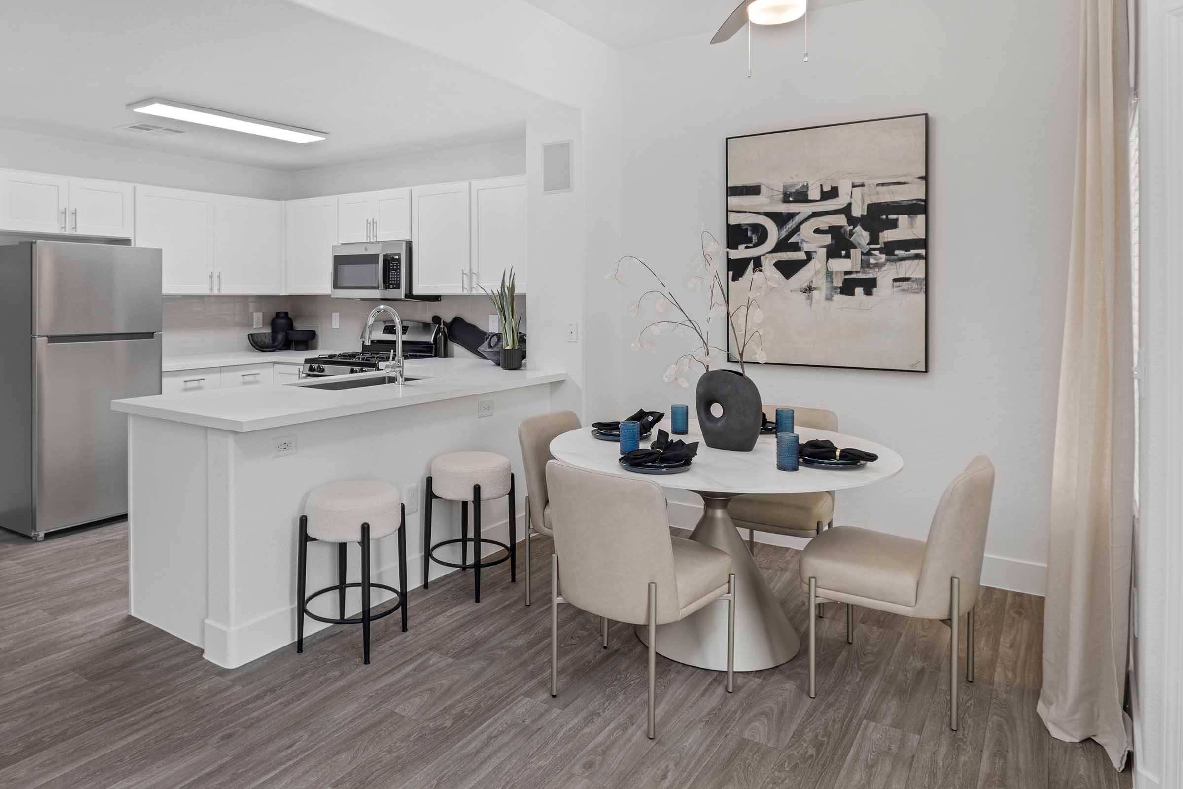 Modern kitchen and dining area featuring a white countertop with seating, stainless steel appliances, and a round dining table set for four. The decor includes a black and white abstract artwork on the wall and stylish beige chairs, creating a sleek and inviting atmosphere.