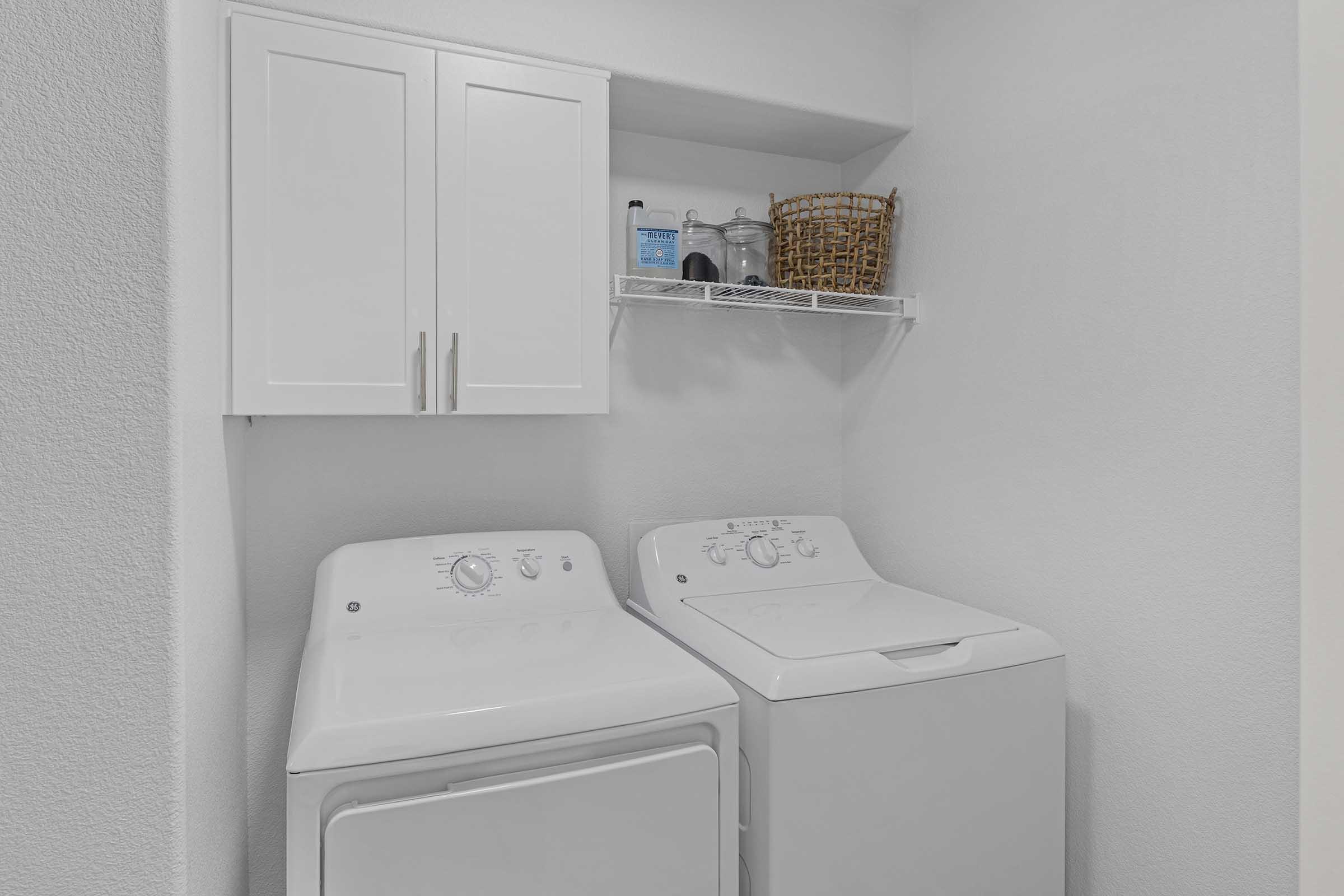 A clean and organized laundry room featuring a white washing machine and dryer side by side. Above, there is a white cabinet with closed doors and a wire shelf holding a basket and cleaning supplies. The walls are painted in a light color, creating a bright and spacious feel.