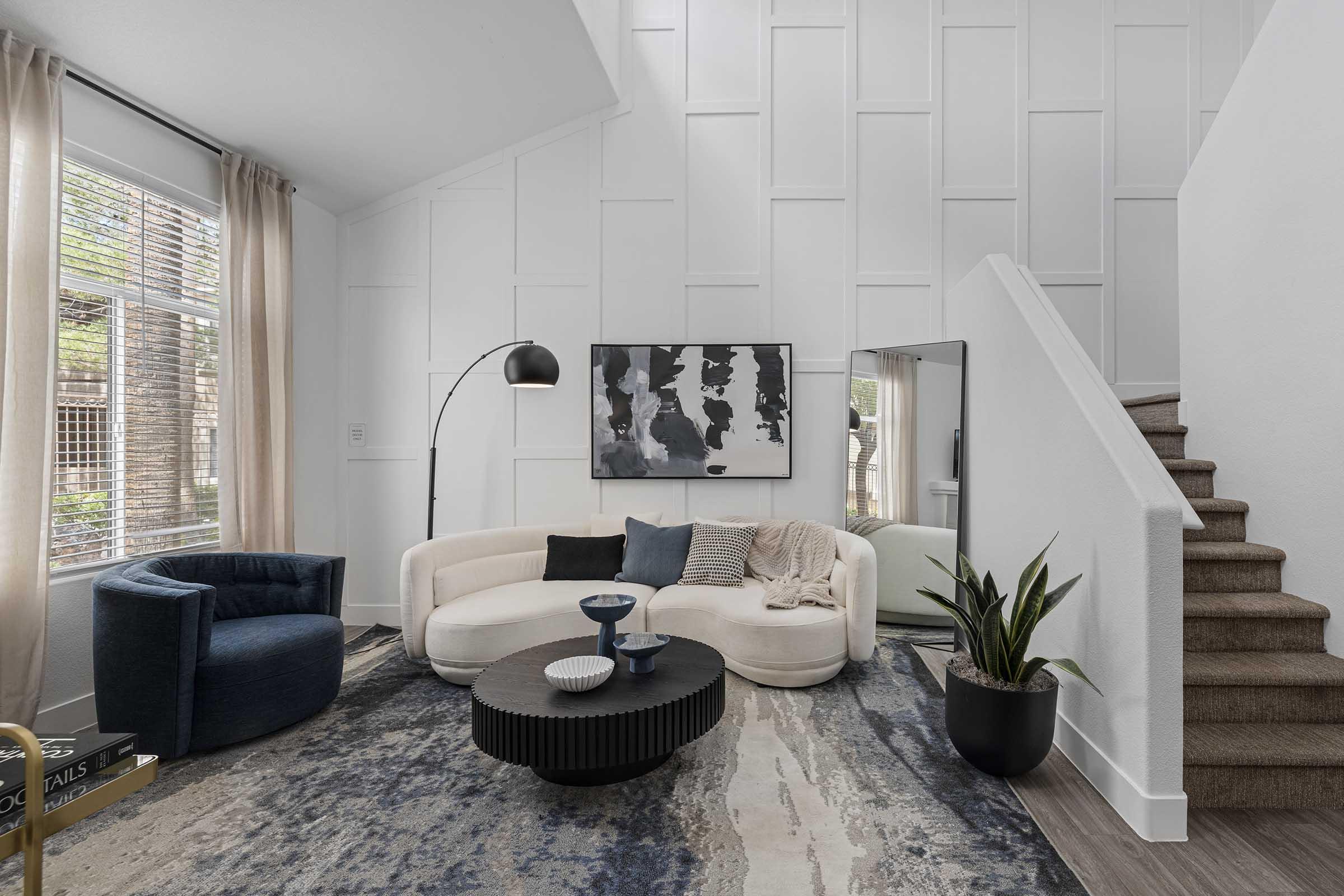 A modern living room featuring a curved white sofa, a round black coffee table, and a dark blue accent chair. The space has large windows with sheer curtains, a decorative wall art piece, and a houseplant. Stairs lead to an upper level, complementing the contemporary design with light colors and textures.
