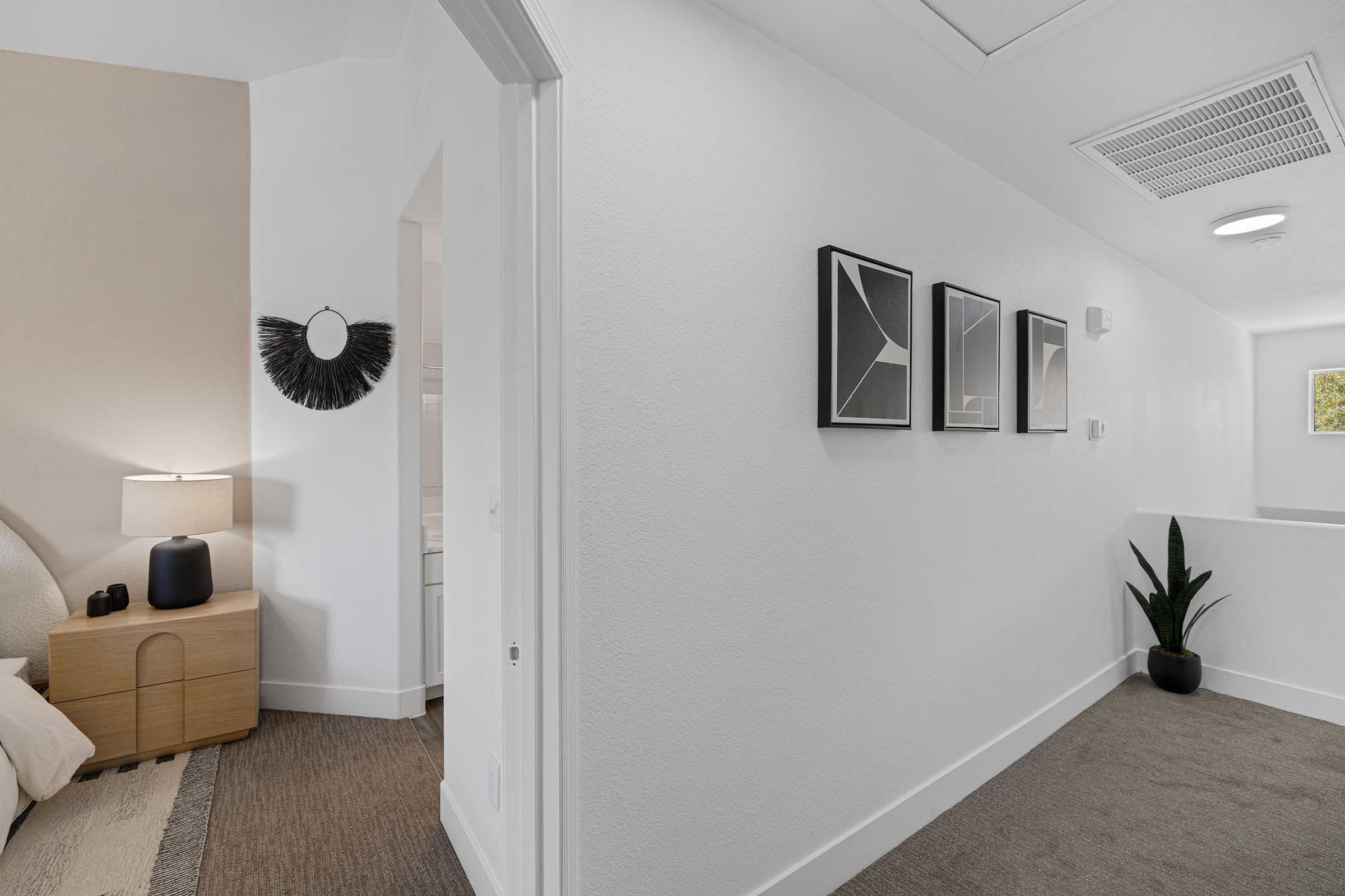 A bright, modern hallway with white walls and light-colored flooring. To the right, there's a small plant near a wall. On the left, a well-decorated room features a lamp and a round mirror. Three framed artworks are arranged on the wall opposite the hallway, contributing to a clean and minimalist aesthetic.