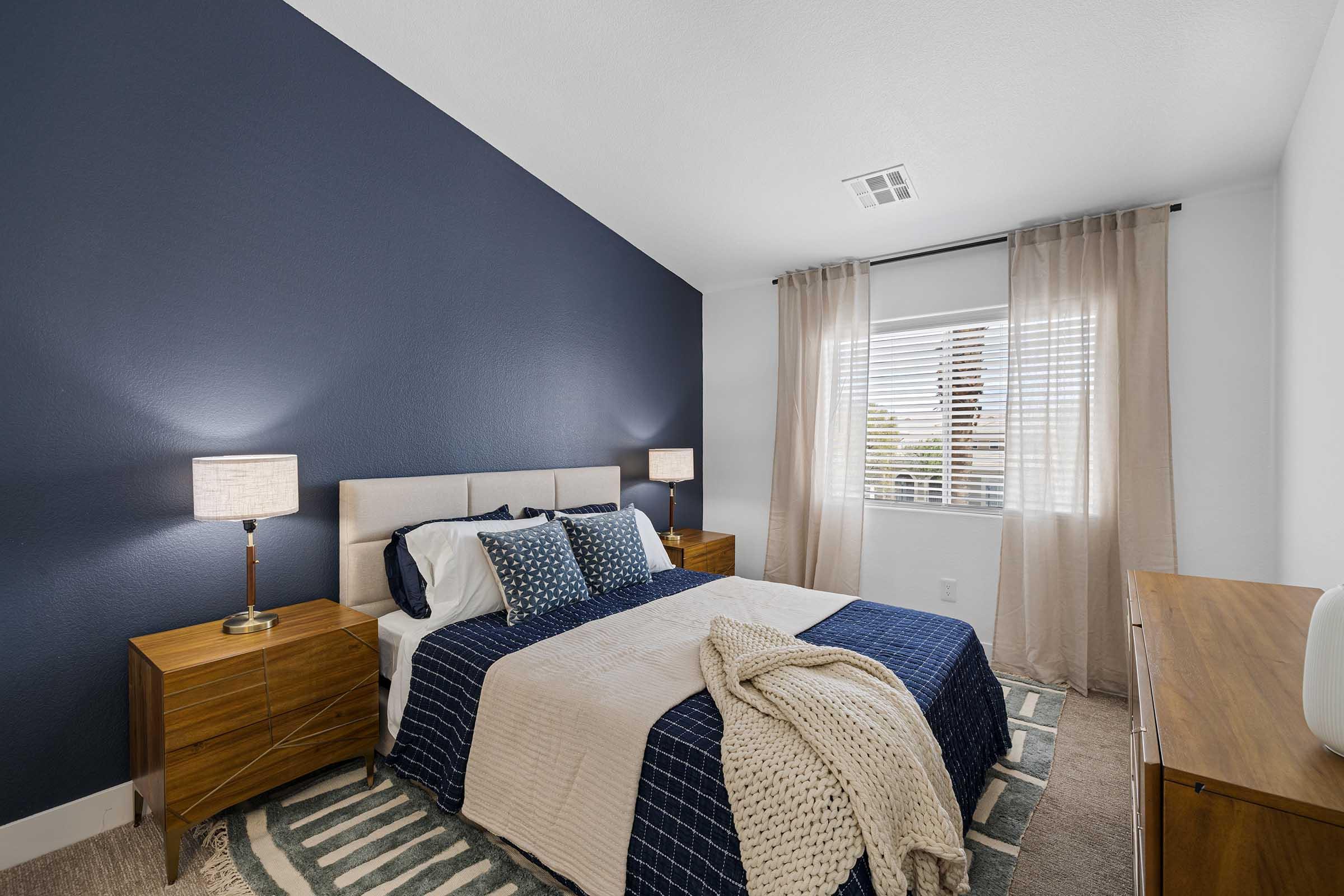 A cozy bedroom featuring a queen-sized bed with navy blue and cream bedding, complemented by decorative pillows. The room has two wooden nightstands with lamps, a large window with sheer curtains, and a soft area rug on the floor, creating a warm and inviting atmosphere.