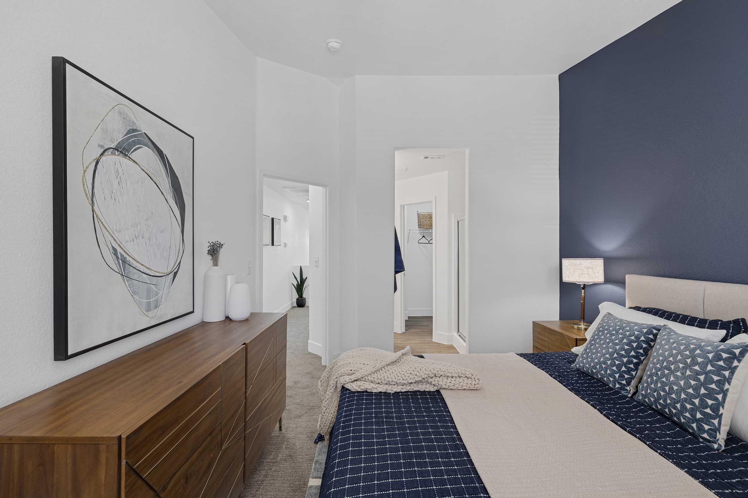A stylish bedroom featuring a wooden dresser, a bed with decorative pillows and a blanket, and a bedside lamp. The wall behind the bed is painted dark blue, while another wall displays an abstract artwork. A doorway leads to another room, creating an inviting and modern atmosphere.