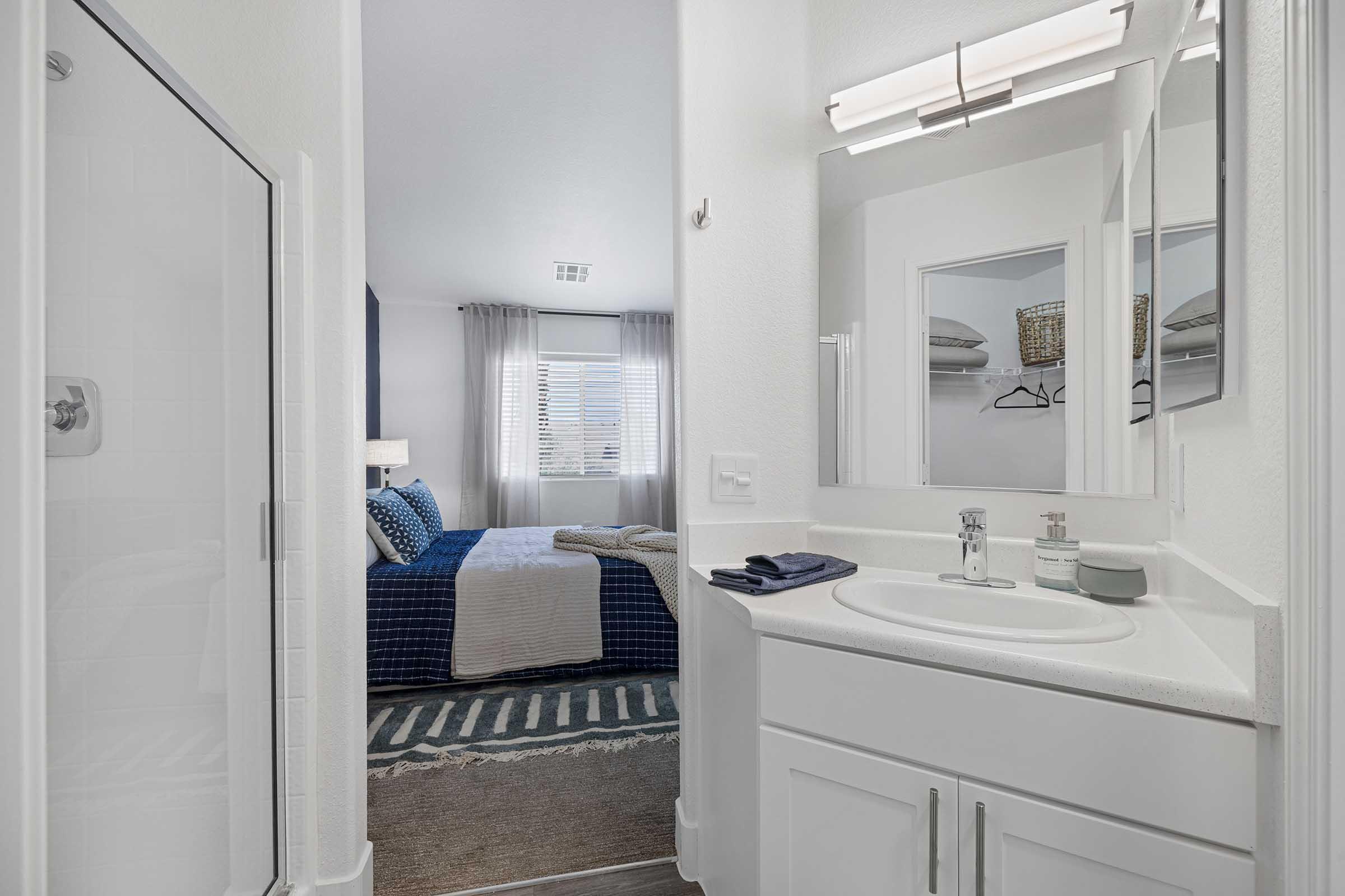 A bright, modern bathroom featuring a shower on the left, a white sink with a countertop, and a mirror above it. In the background, a cozy bedroom is visible through an open door, showcasing a blue and white bedding set and large windows with curtains.