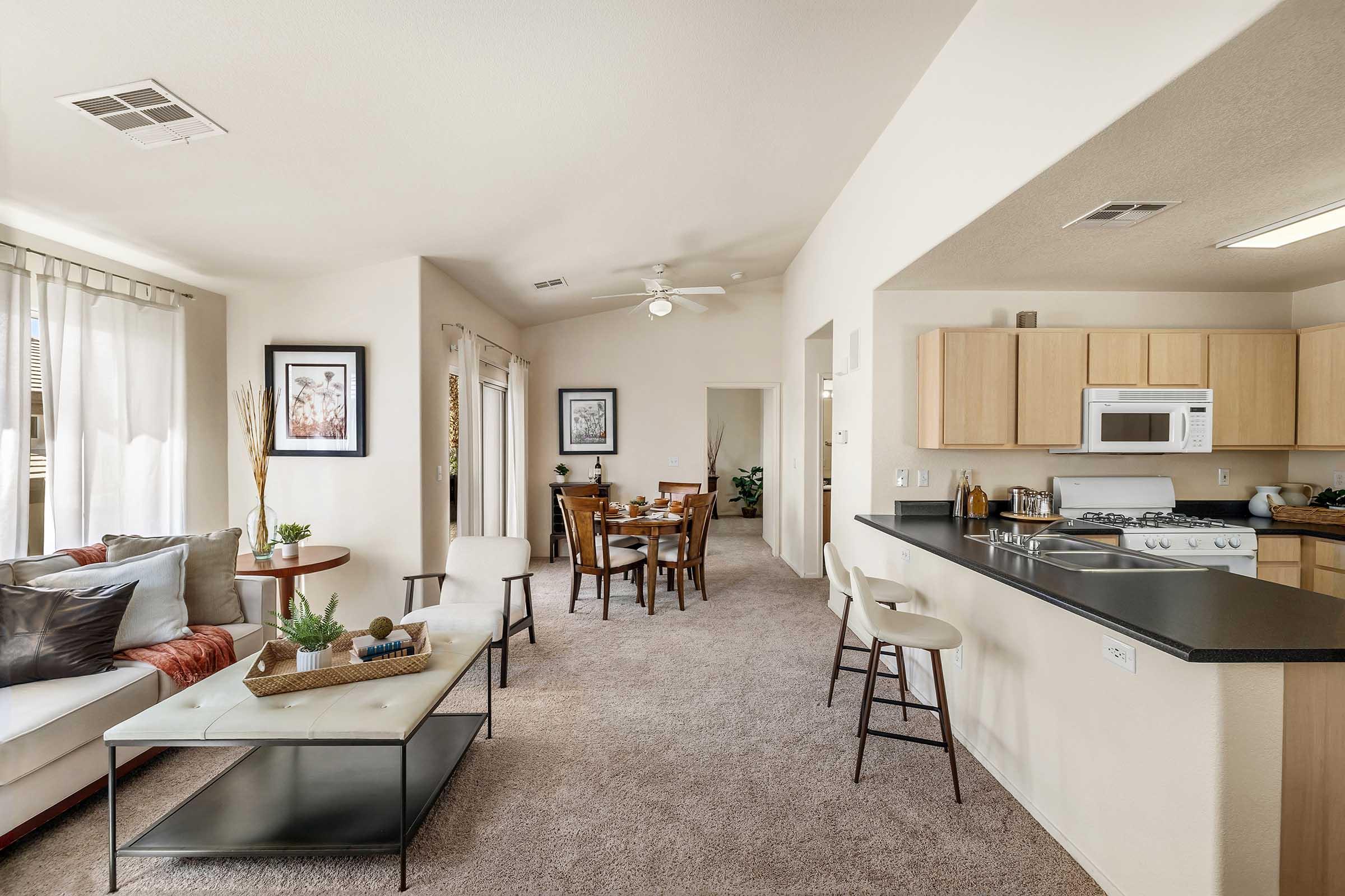 A bright and spacious living area featuring a beige couch, a coffee table, and a dining table with chairs. The kitchen is open with light wood cabinetry. Large windows allow natural light to fill the room, which has neutral-colored walls and carpeting, creating a cozy atmosphere.