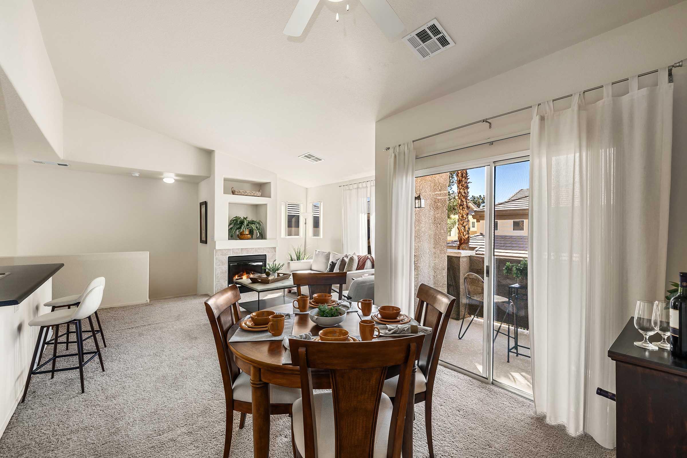 A spacious living and dining area featuring a round wooden dining table set for four, a cozy seating arrangement with a couch and armchair, and large sliding doors leading to an outdoor patio. The room has light-colored walls, neutral carpeting, and large windows allowing natural light to fill the space.