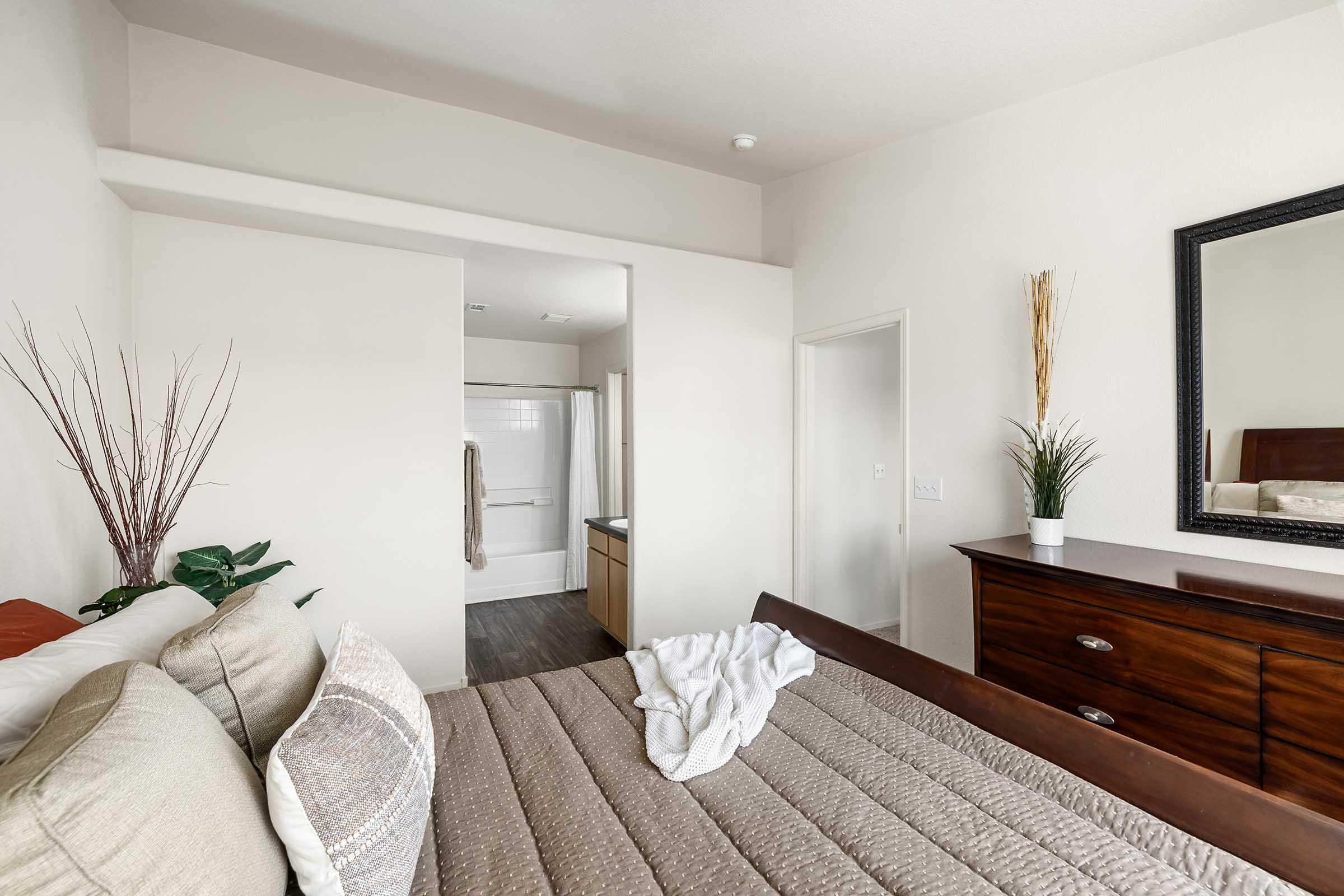 A cozy bedroom featuring a wooden bed with a neatly made quilt and pillows. A decorative plant is placed by the bed, and a large mirror hangs on the wall. In the background, there is a small bathroom visible, with a shower and vanity area. The overall color palette is neutral and calming.