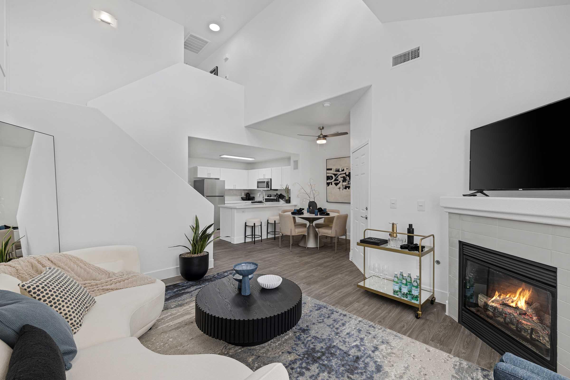 A bright and modern living room featuring a white sectional sofa, a round black coffee table, and a sleek fireplace. In the background, there's a dining area with beige chairs and a kitchen with contemporary appliances. Large windows let in natural light, enhancing the open, airy feel of the space.