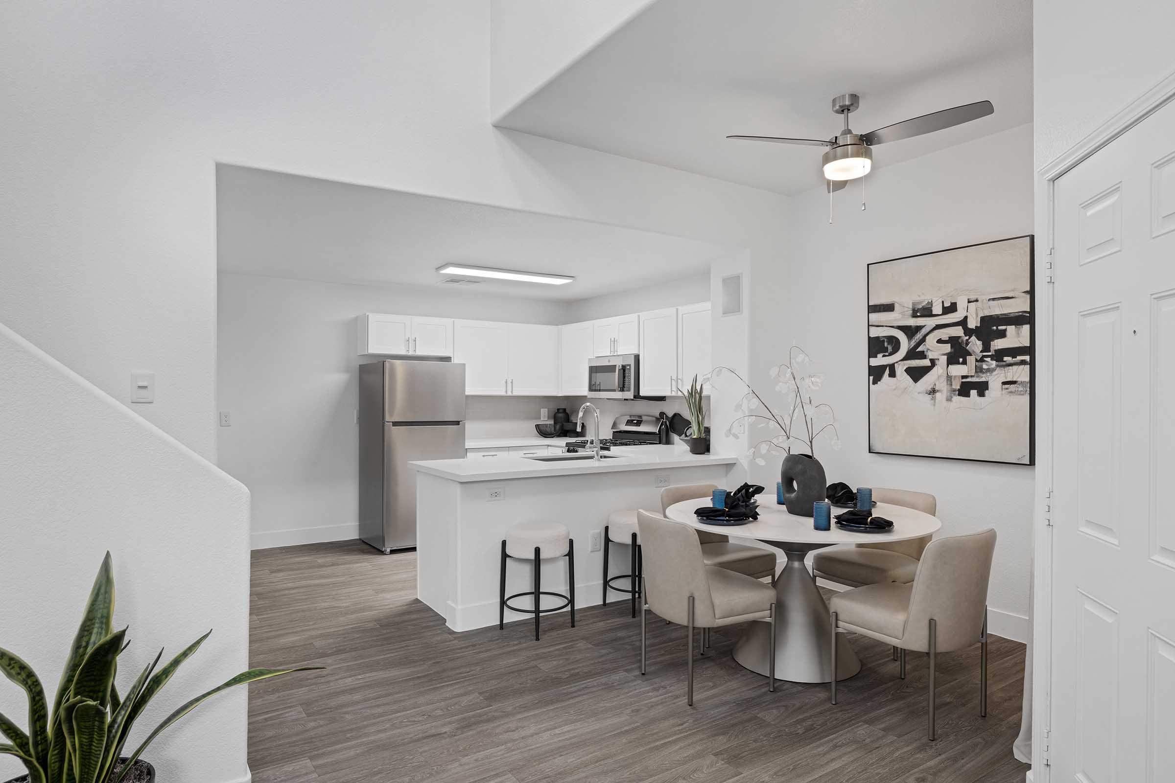 A modern kitchen and dining area featuring white cabinetry, stainless steel appliances, and a round dining table with beige chairs. A large painting decorates the wall, and a plant adds a touch of greenery. The space is well-lit and has a clean, contemporary design with hardwood flooring.