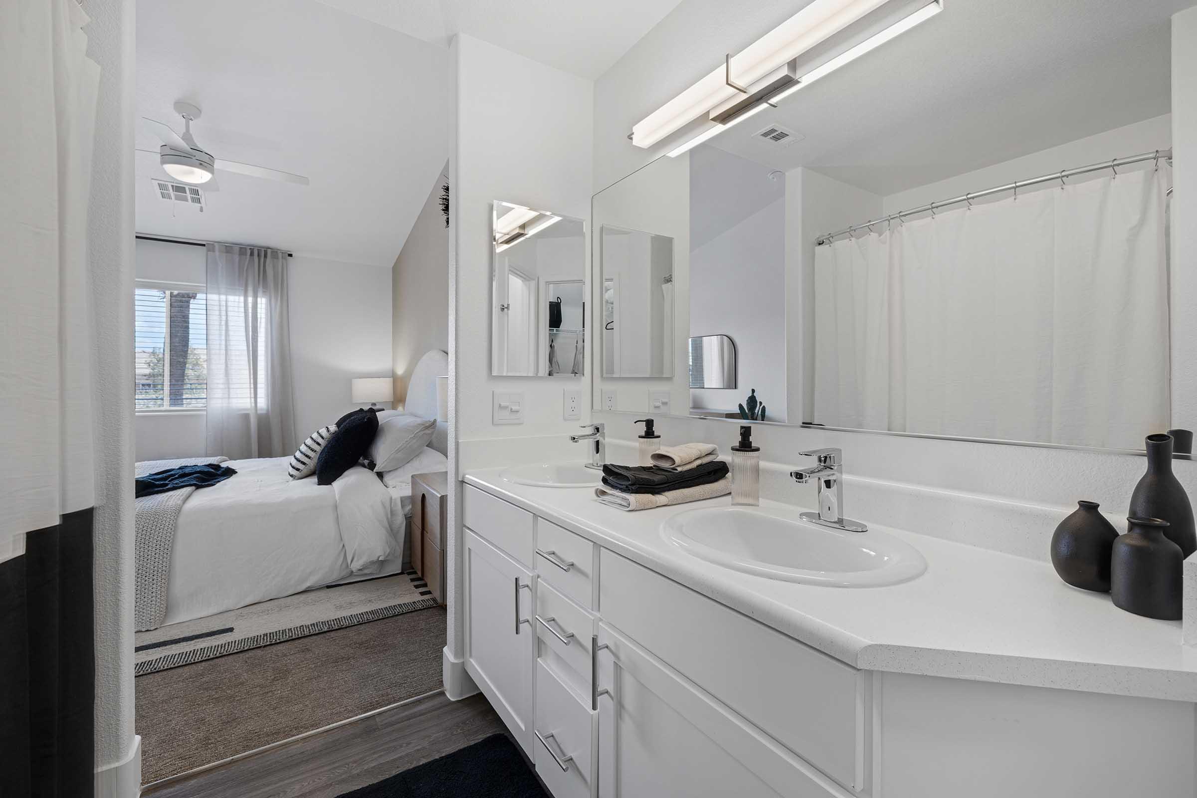 A modern bathroom featuring a white countertop with a sink, neatly arranged towels, and decorative vases. In the background, there is a view of a bedroom with a bed dressed in white linens and a blue throw blanket. Natural light streams in through a window, enhancing the airy atmosphere.