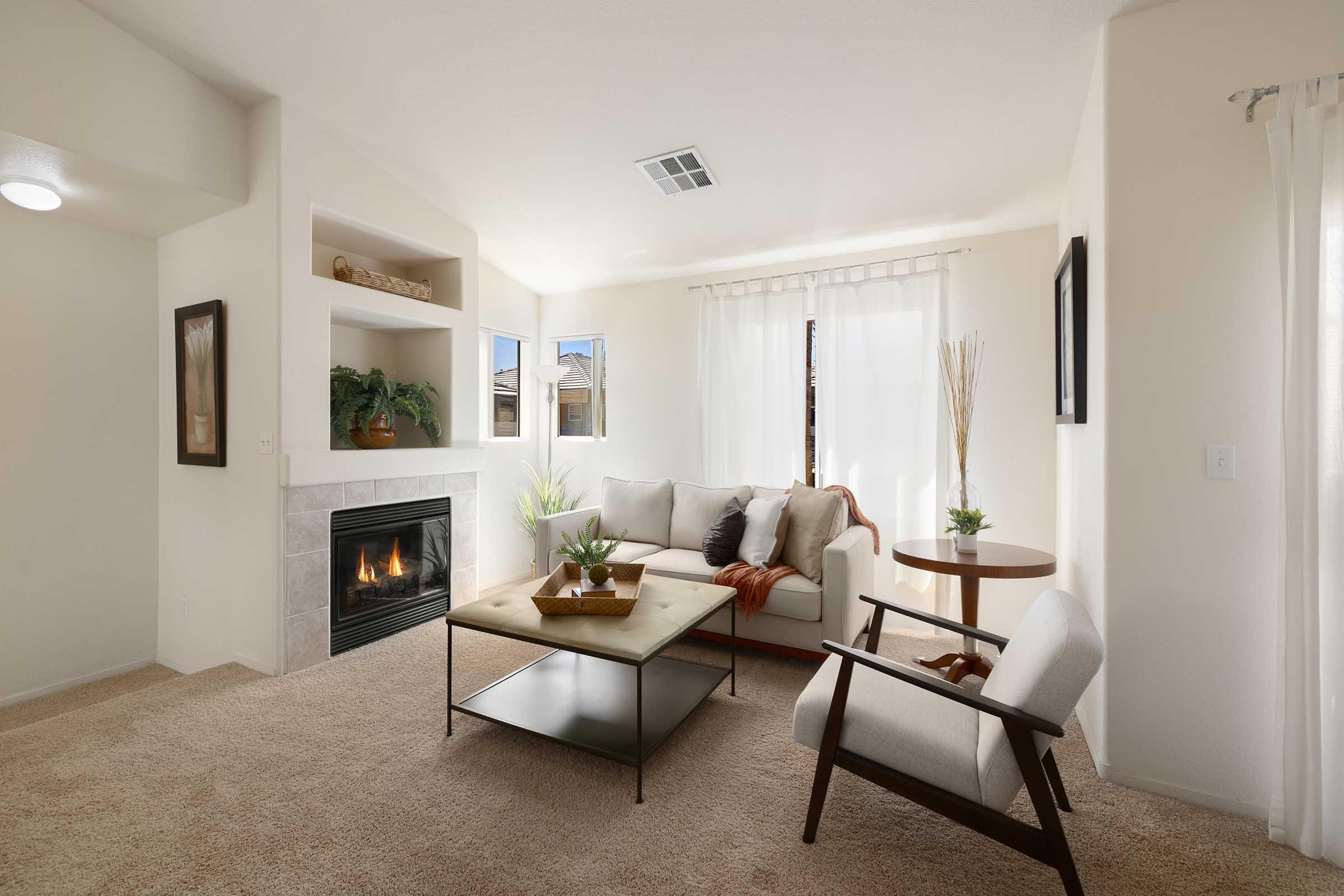 Modern living room featuring a cozy seating area with a white sofa adorned with cushions, a coffee table at the center, and an armchair. A fireplace is built into the wall, surrounded by decorative plants and wall art. Natural light streams through large windows, creating a warm and inviting atmosphere.