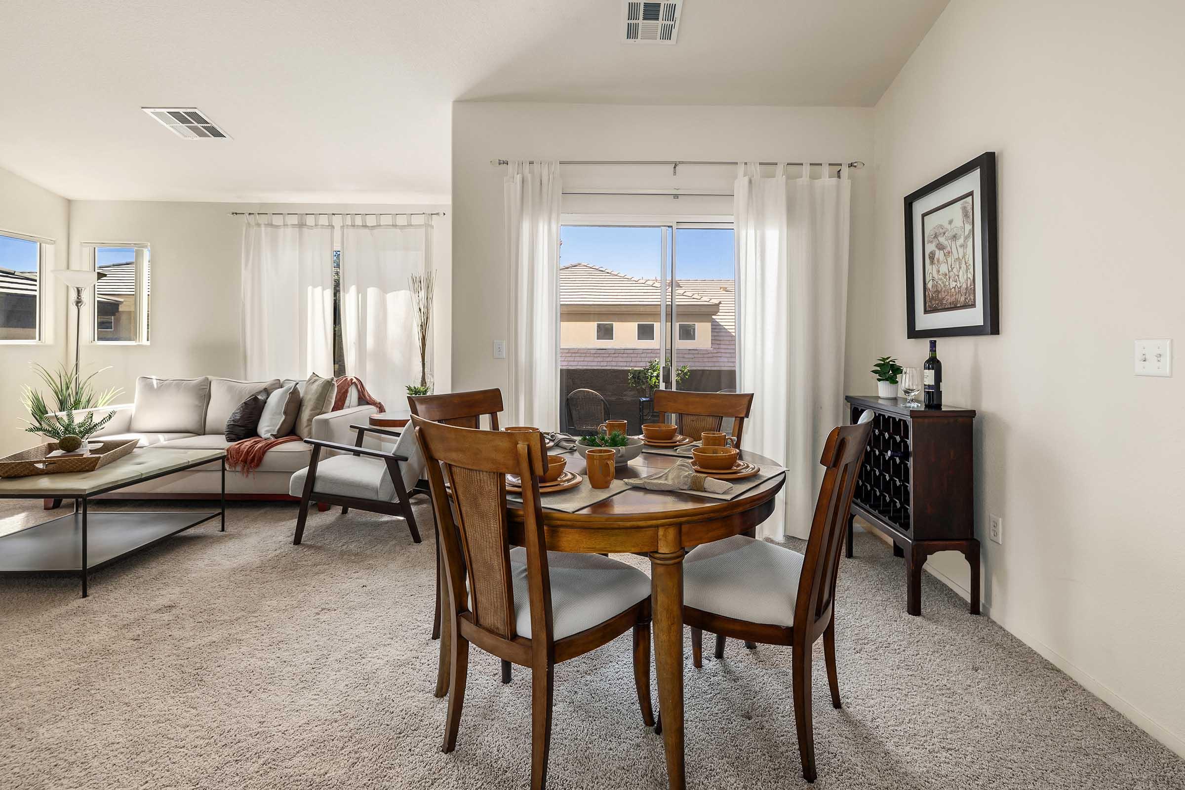 A cozy dining area featuring a round wooden table set for four, surrounded by upholstered chairs. In the background, a light-filled living space with a sectional sofa, decorative pillows, and a window with sheer curtains. A cabinet holds bottles, and plants provide a touch of greenery.