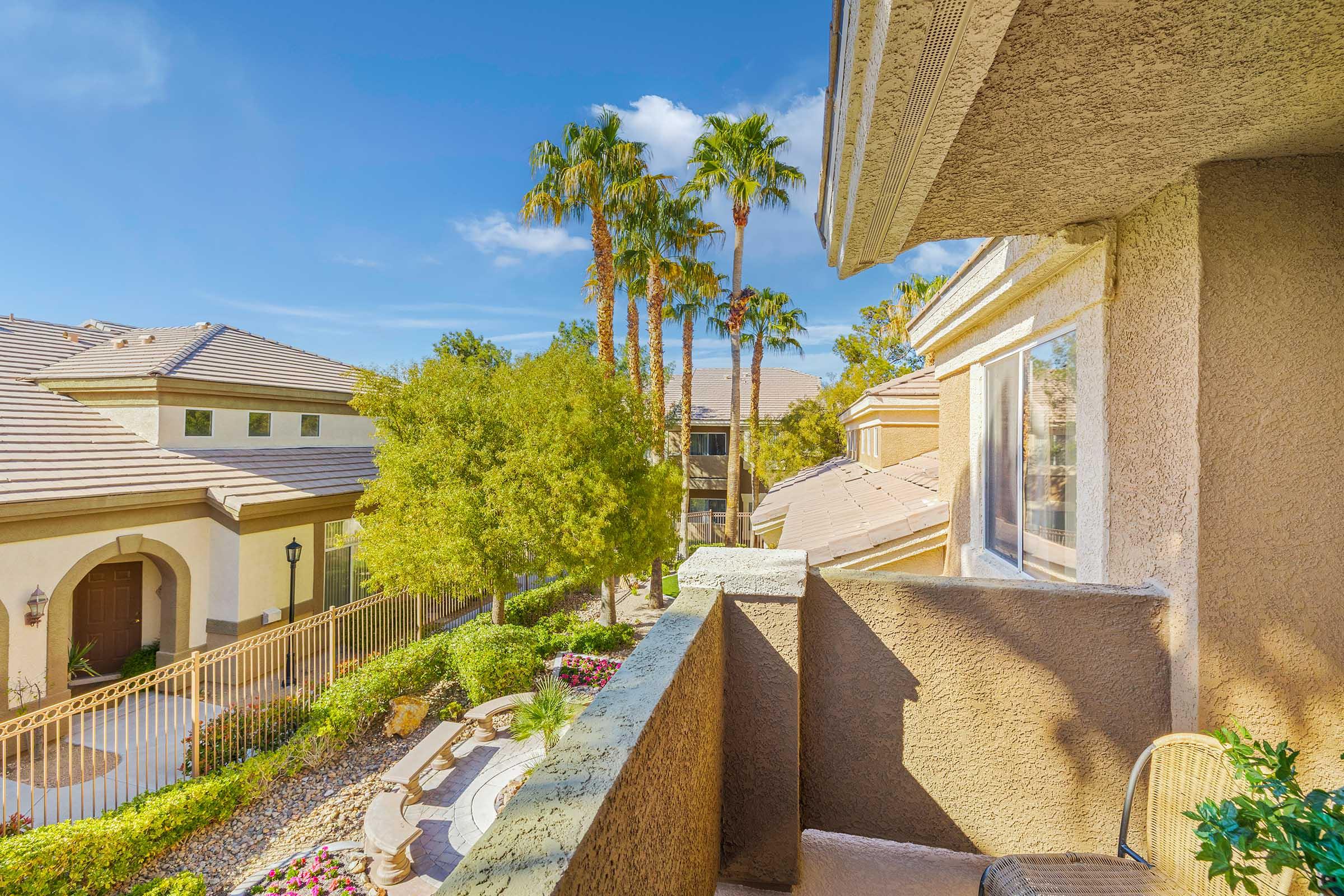 A scenic view from a balcony featuring palm trees, well-maintained landscaping, and residential buildings under a clear blue sky. The setting suggests a warm, sunny climate, with colorful flowers and pathways visible in the foreground.