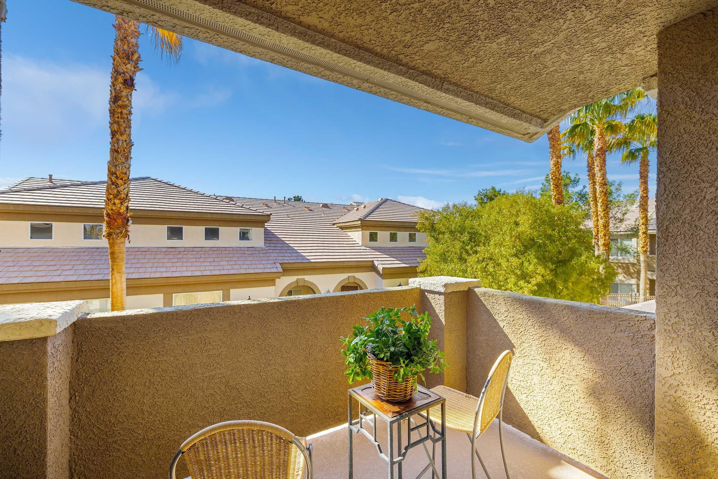 A cozy balcony view featuring a small table with a potted plant and two chairs. The scene includes palm trees in the background, and neighboring rooftops under a clear blue sky, creating a serene and inviting outdoor space.