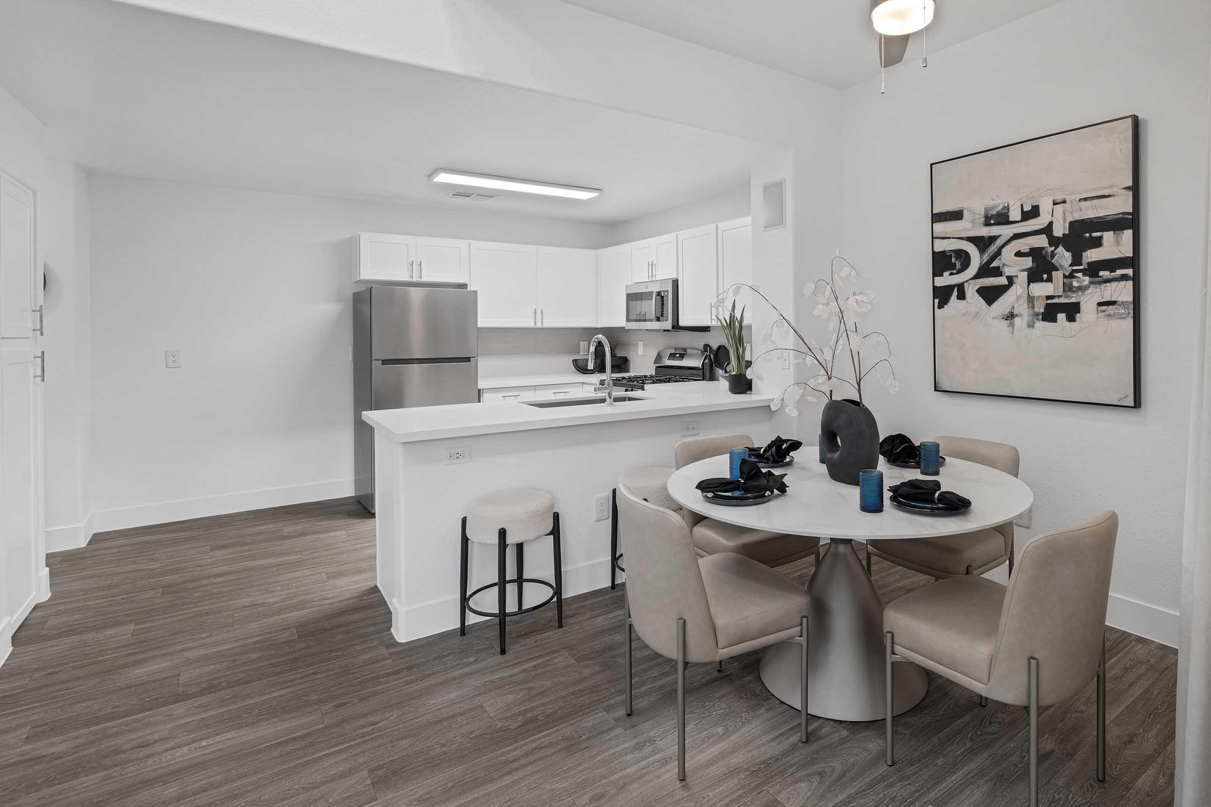 A modern kitchen and dining area featuring a round white table with black dinnerware, surrounded by beige upholstered chairs. The kitchen includes white cabinets, stainless steel appliances, and a sleek design. A piece of abstract artwork hangs on the wall, adding a touch of elegance to the space.