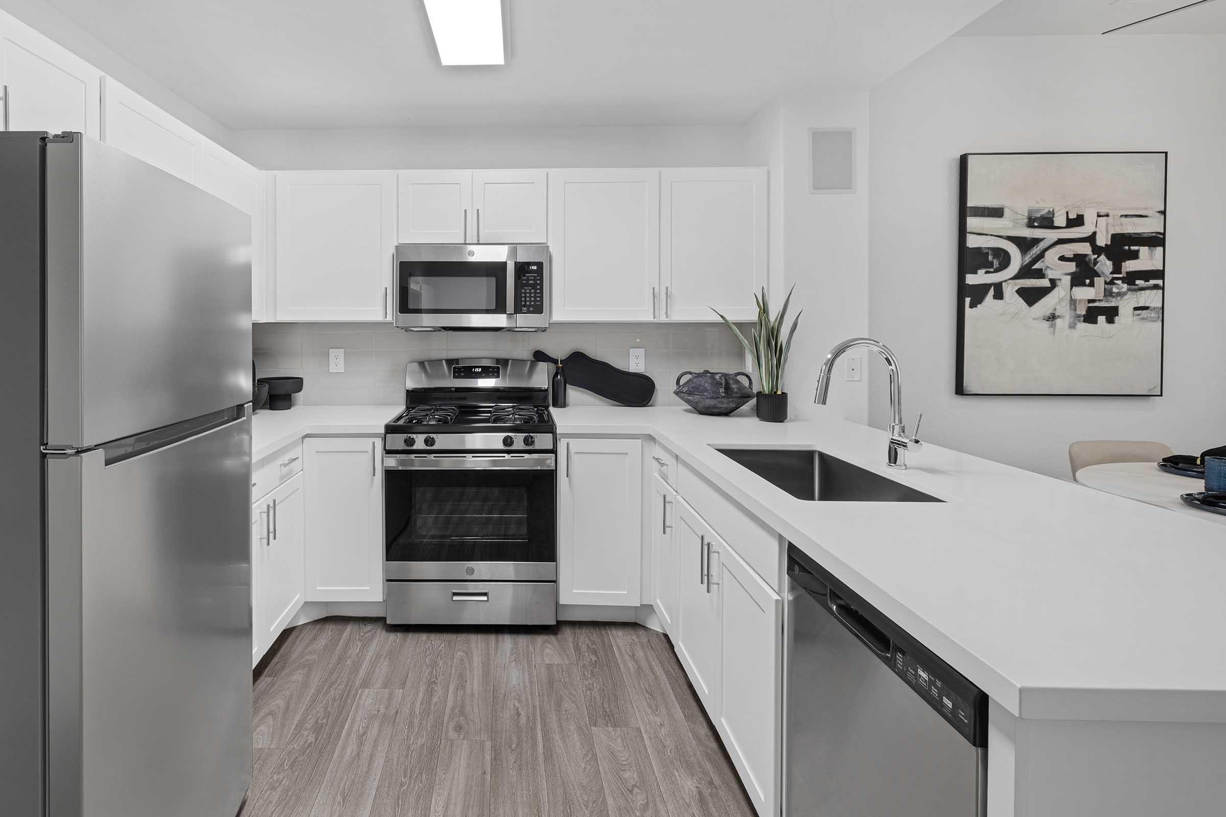 Modern kitchen featuring stainless steel appliances including a refrigerator, microwave, and gas stove, white cabinets, and a sleek countertop. The space has a minimalist design with wood flooring and decorative elements like a plant and artwork on the wall.