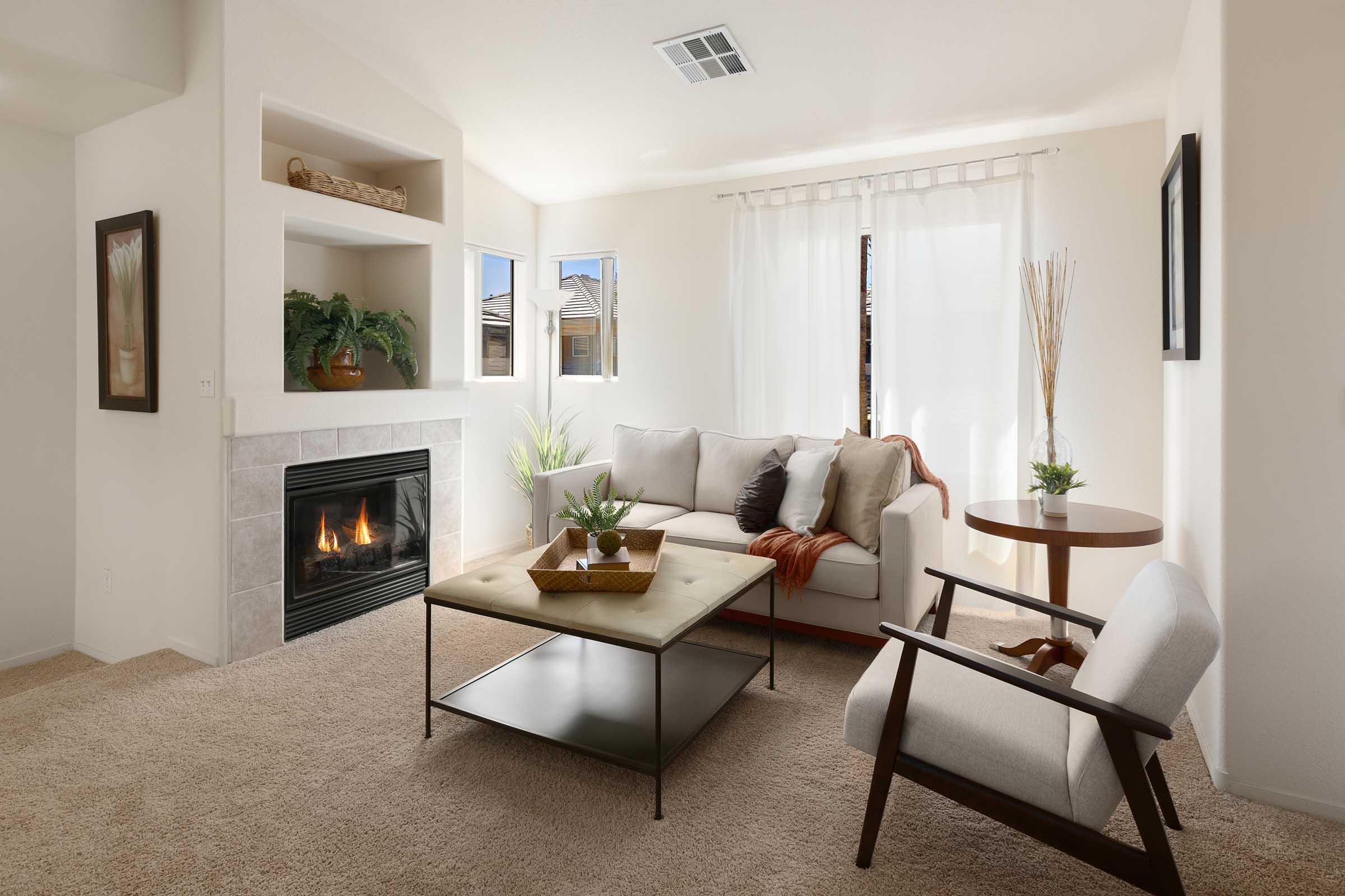 A cozy living room featuring a beige sofa with decorative pillows, a wooden coffee table on a light carpet, an armchair, a fireplace with a stone surround, and natural light coming through window treatments. Decorative plants and a small side table enhance the inviting atmosphere.
