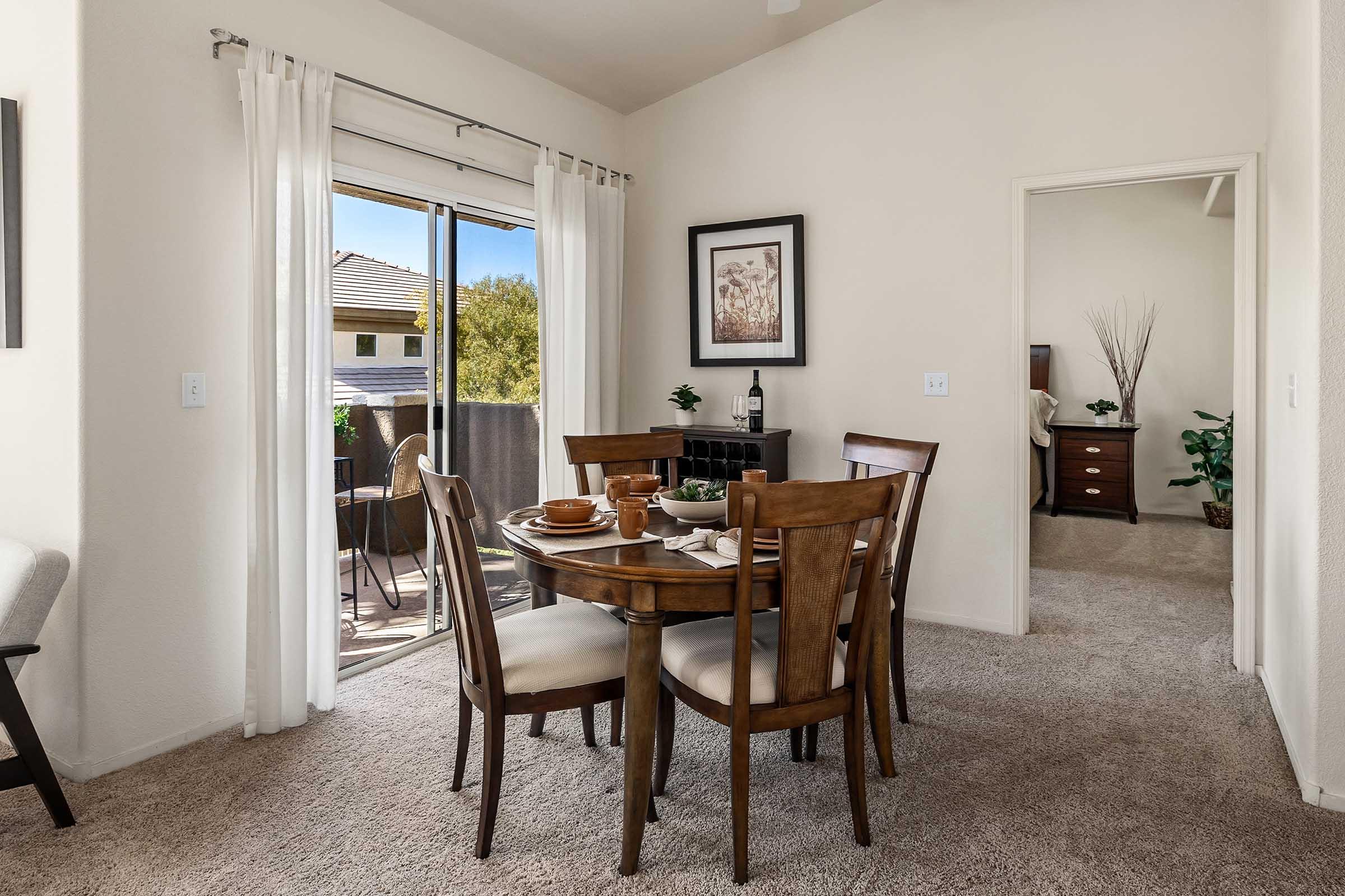 A cozy dining area featuring a round wooden table set for a meal, with four chairs. Floor-to-ceiling windows allow natural light in, leading to an outdoor balcony. Soft beige walls and light carpet create a warm ambiance. A door leads to another room, and decorative plants add a touch of greenery.